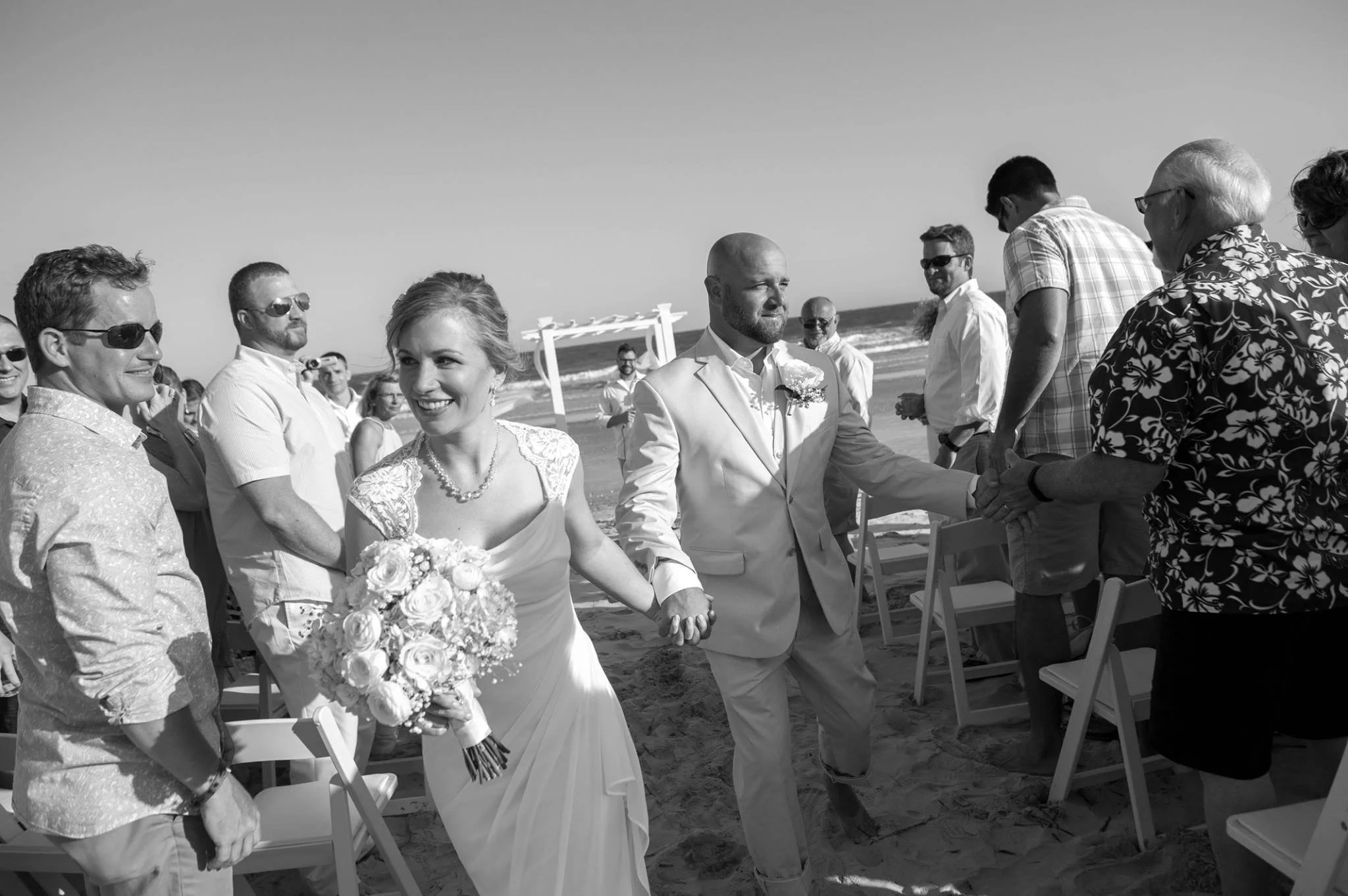 Black and white photo of a beach wedding with a bride and groom holding hands, surrounded by friends and family, with an ocean backdrop.