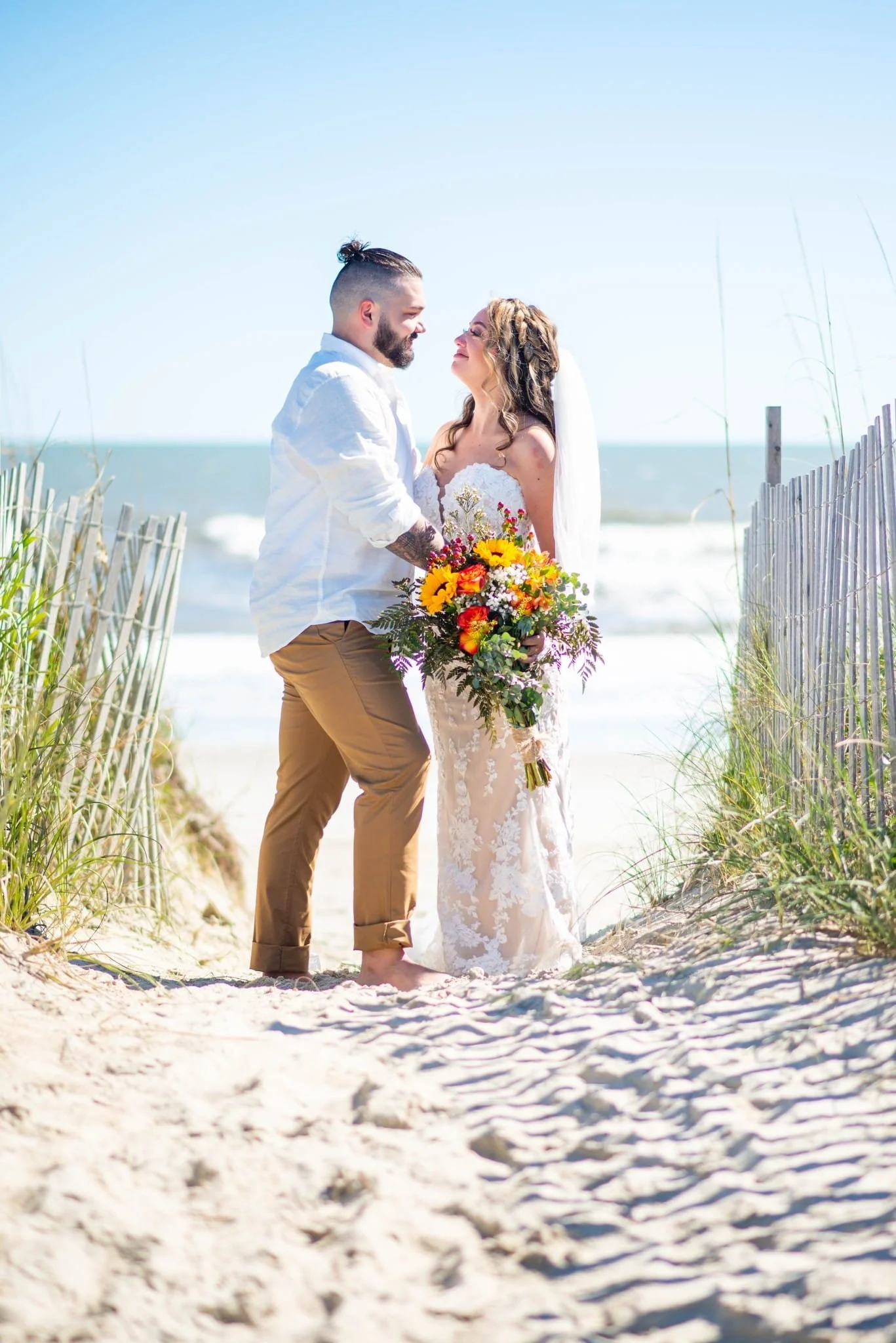 A newlywed couple standing on a beach, holding hands, with the ocean and sky in the background. The bride is in a lace wedding dress holding a colorful bouquet, and the groom is in a white shirt and tan pants.