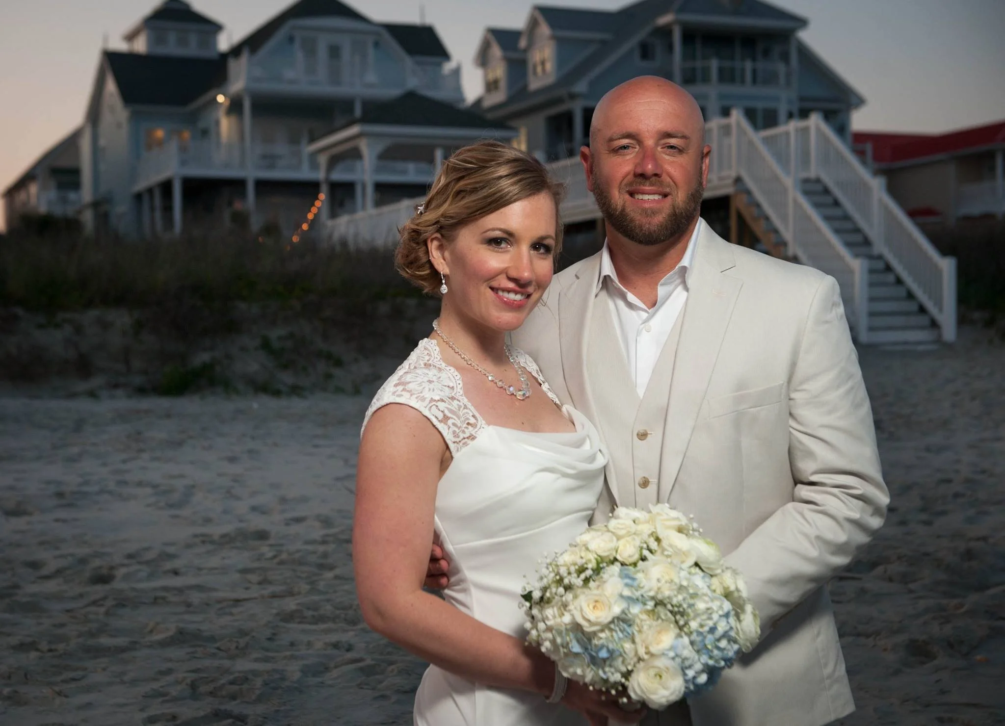 A newlywed couple in wedding attire standing on a beach at sunset, holding a bouquet of white flowers, with houses in the background.