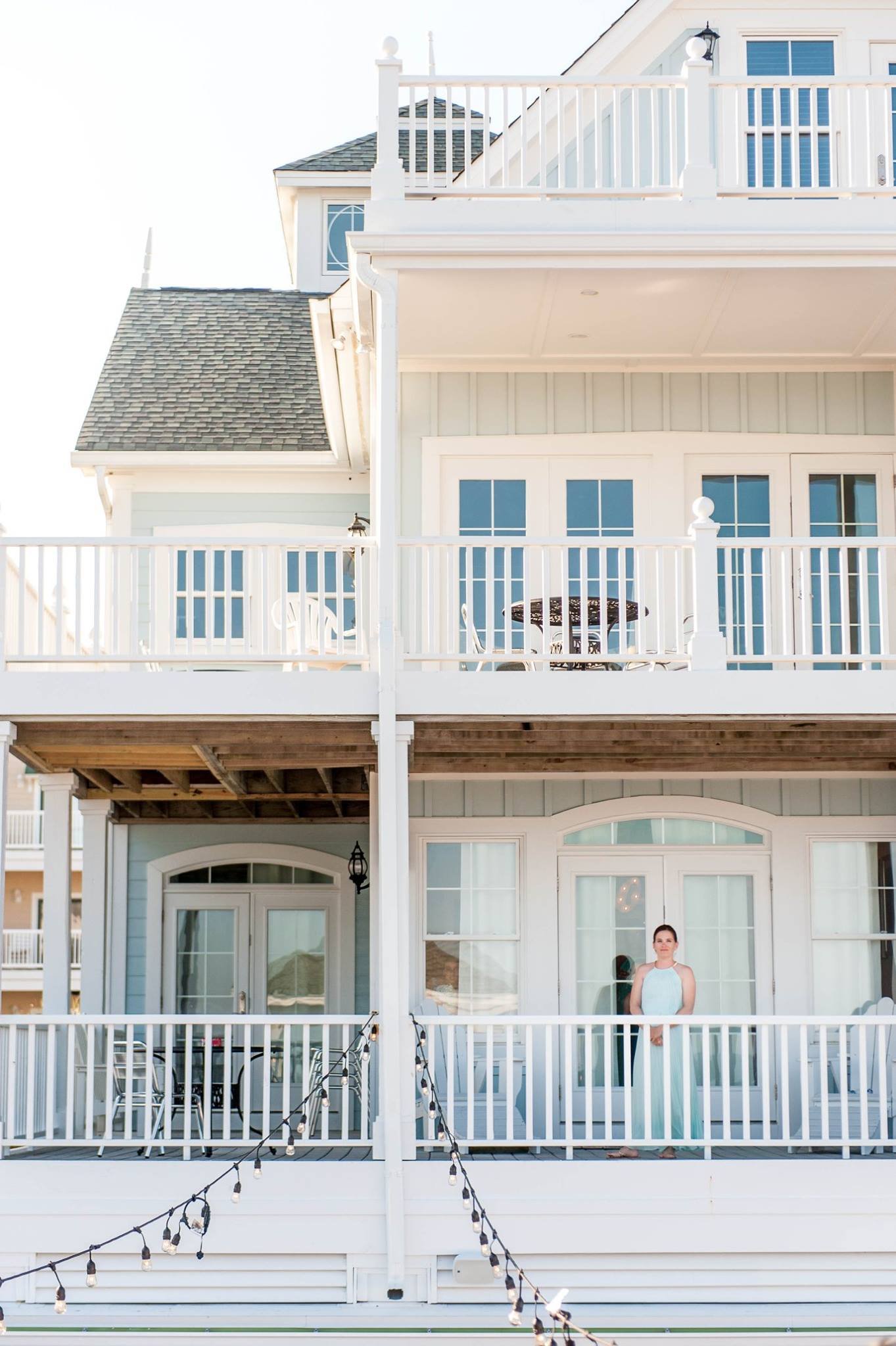 A woman standing on the balcony of a multi-story seaside house with white railings and string lights hanging in the foreground.
