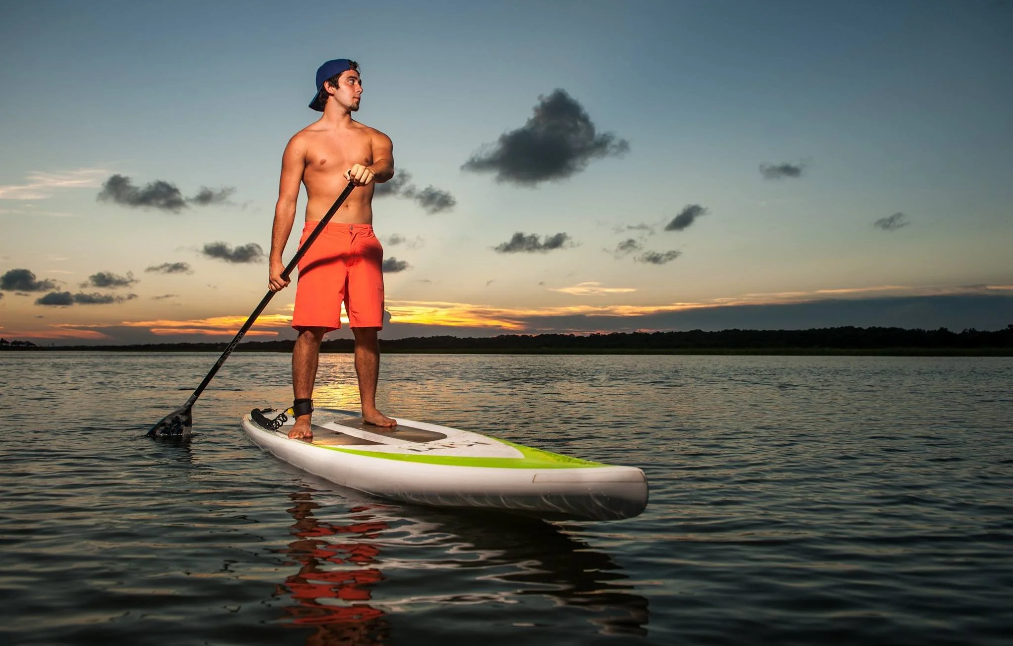 A young man paddleboarding on a lake at sunset, wearing red swim shorts and a blue cap, holding a paddle, with a cloudy sky in the background.