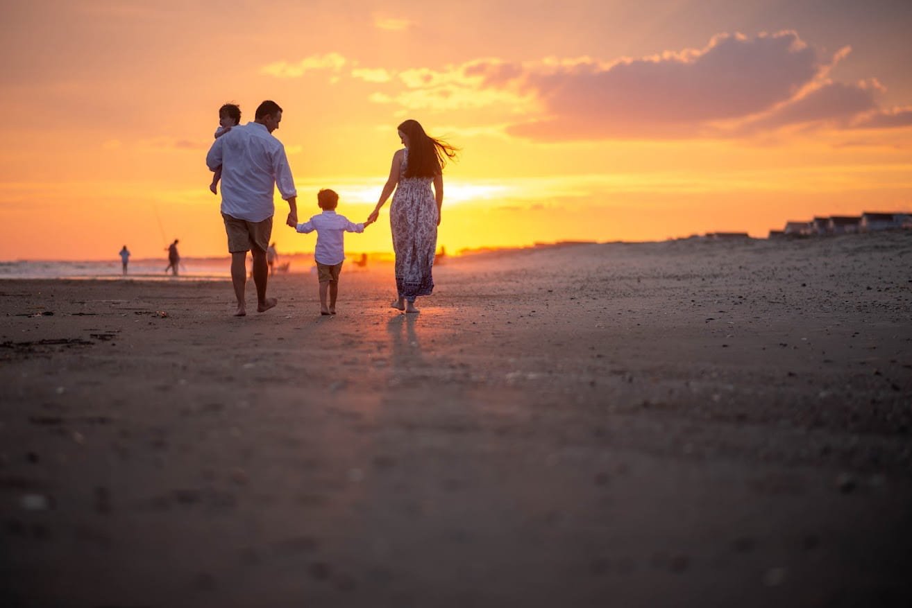 Family walking hand in hand on the beach during sunset, with a father, mother, and two children, all silhouetted against the colorful sky.