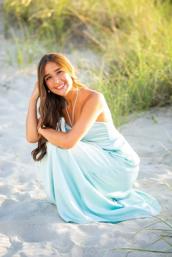 A young woman in a light blue dress sitting on the sandy beach with green grass in the background, smiling at the camera.