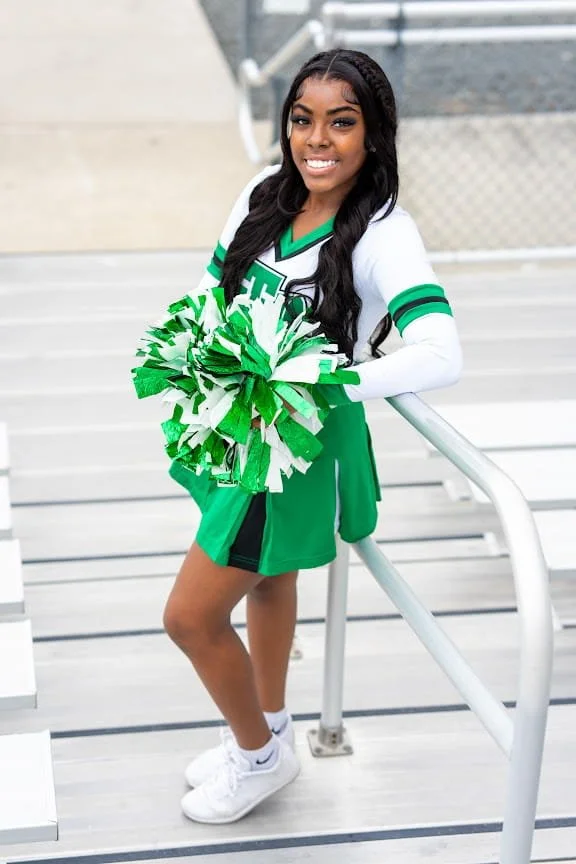 A young woman in a green and white cheerleading uniform, holding green and white pom-poms, standing outdoors on a staircase, smiling at the camera.