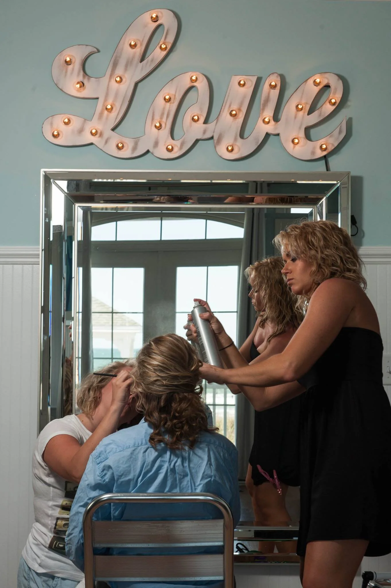 A hairstylist is blow-drying a woman's hair in front of a mirror decorated with a marquee sign that spells out "Love" in bright lights.