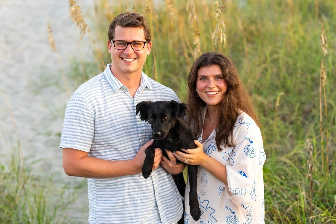 A happy couple with a black dog at the beach with tall grass in the background during sunset.