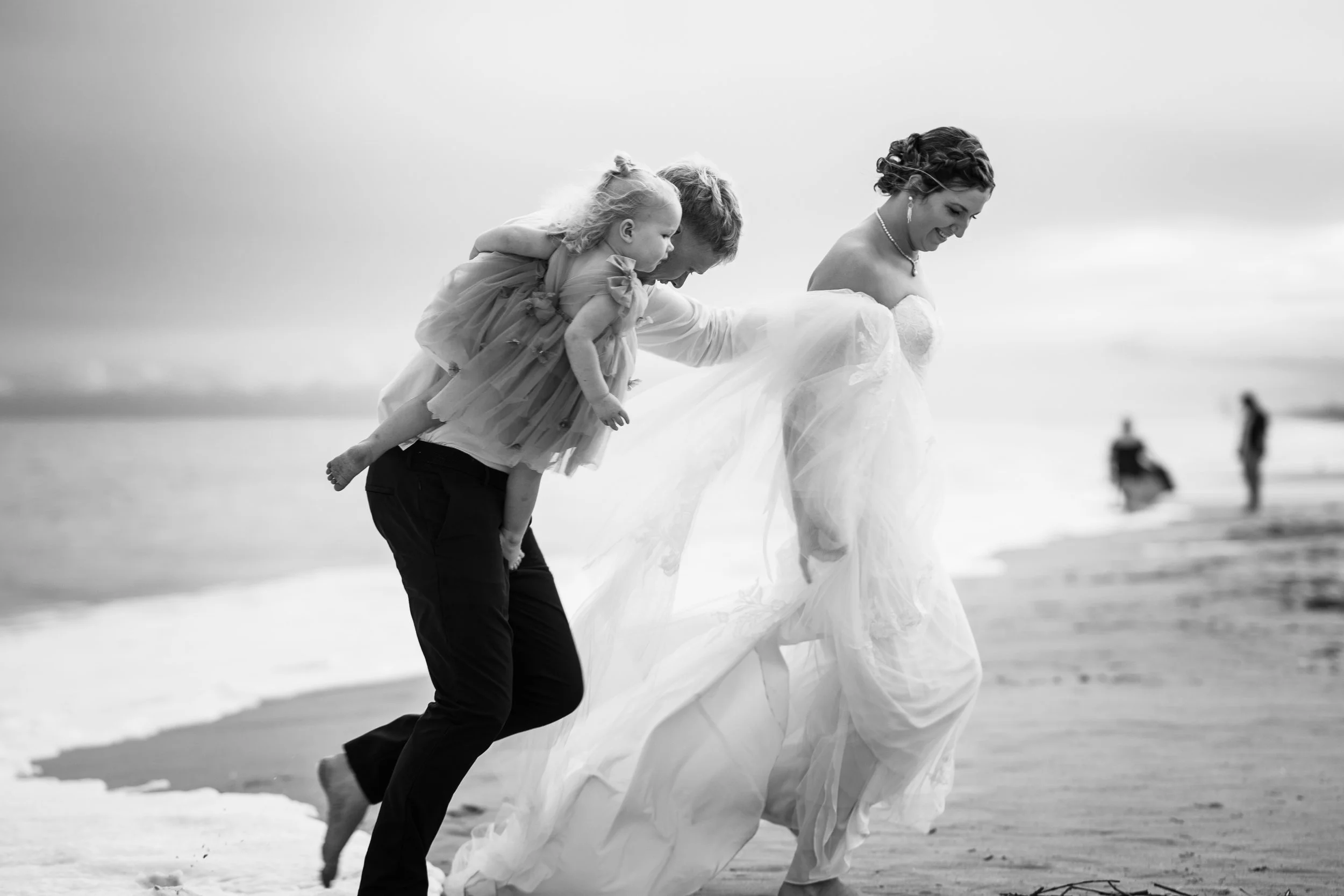 A woman in a wedding dress holding the hand of a man and a child on a beach during daytime, with other people in the distance.