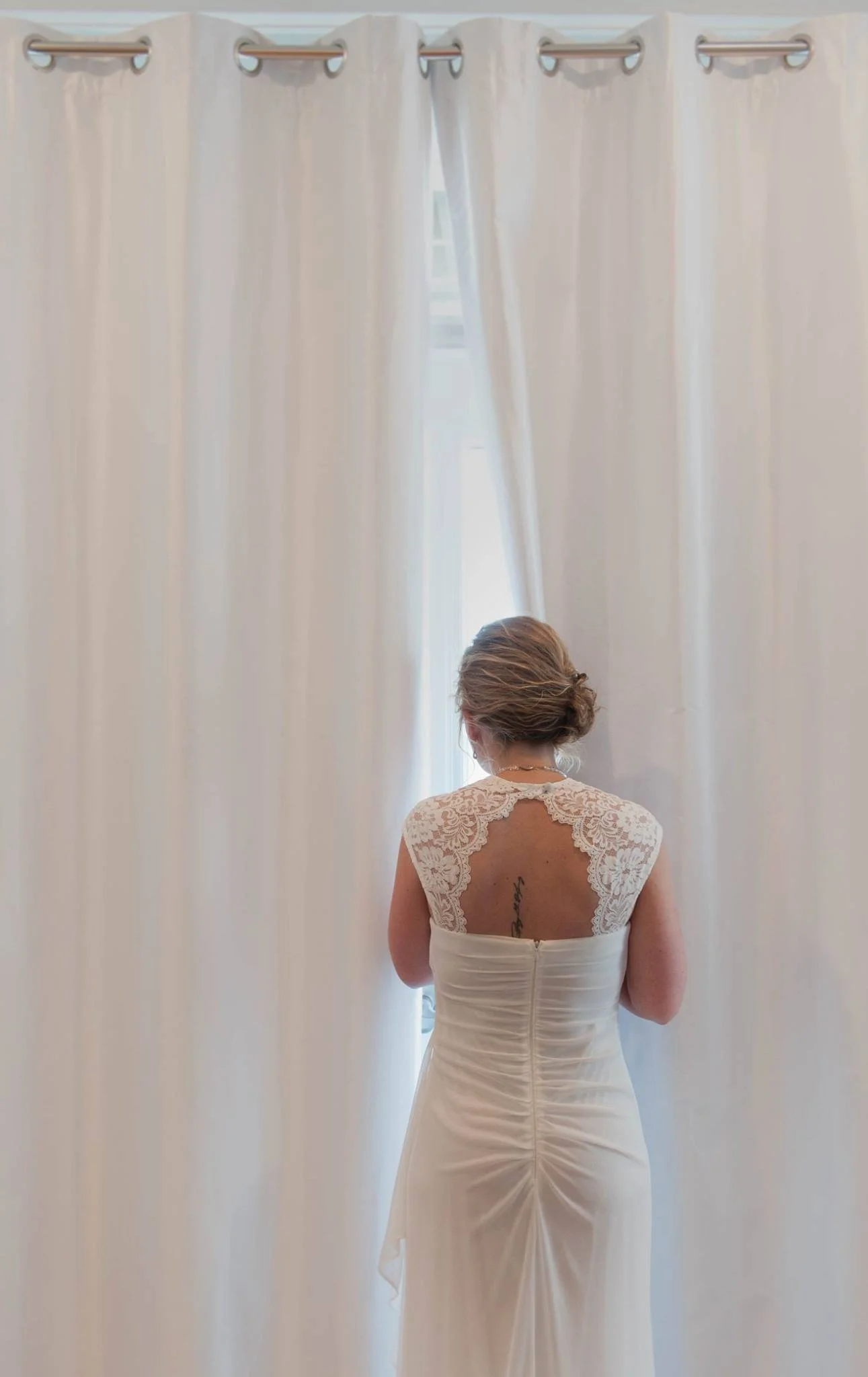 A woman in a white wedding dress with lace details on the back, facing away and looking out of a window through cream-colored curtains.