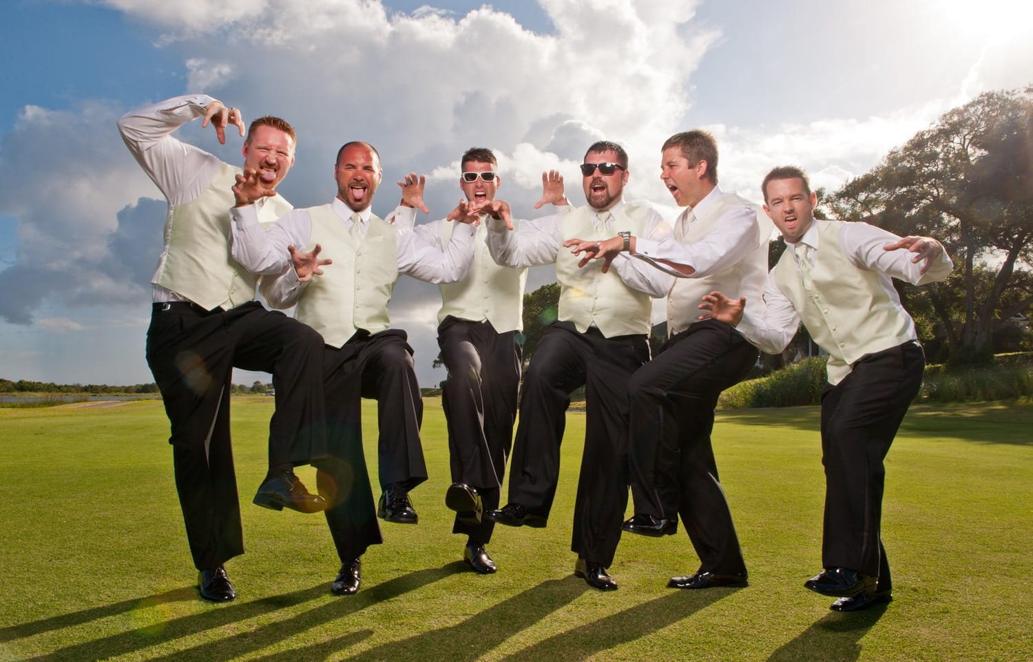 A group of seven men dressed in white shirts, vests, and black pants, posing on a golf course with playful gestures against a cloudy sky and trees in the background.