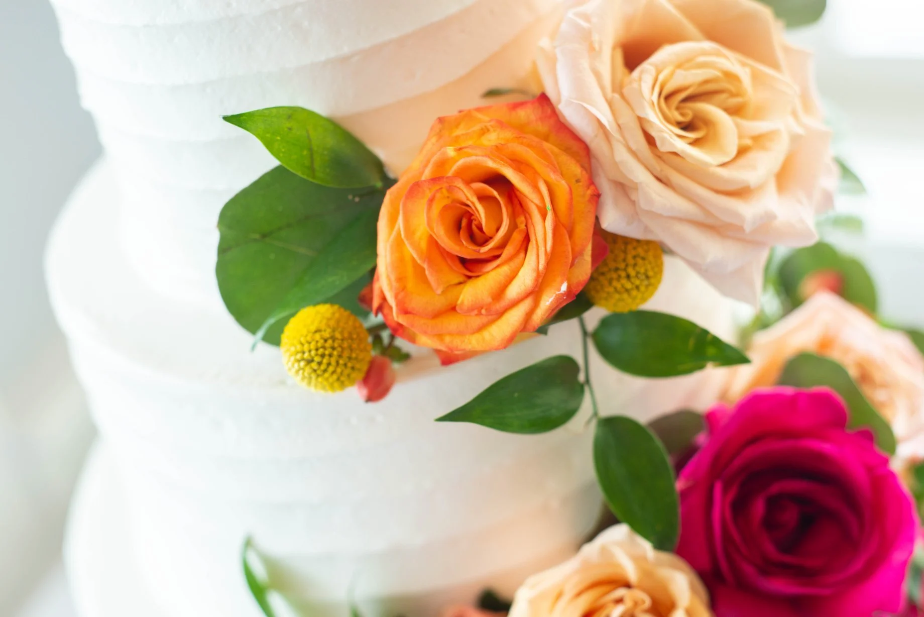 Close-up of a white wedding cake decorated with orange, peach, cream, and pink roses, along with green leaves and small yellow flowers.