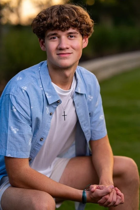 A young man with curly brown hair sitting outdoors on grass, wearing a light blue button-up shirt over a white T-shirt with a cross necklace, smiling slightly.