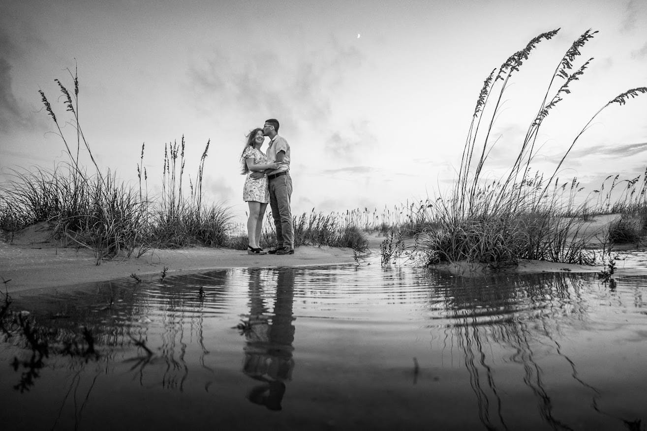 A black-and-white photo of a couple standing on a sandy beach near water, embracing and smiling, with tall grasses and a cloudy sky in the background.