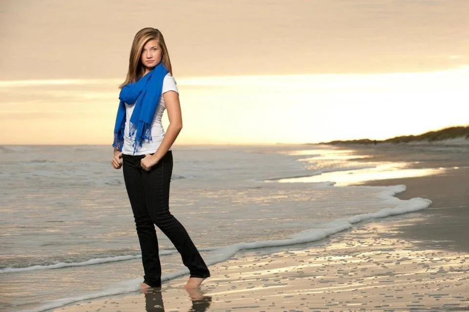 A woman with long blonde hair stands barefoot on the beach at sunset, wearing a white T-shirt, black pants, and a blue scarf.
