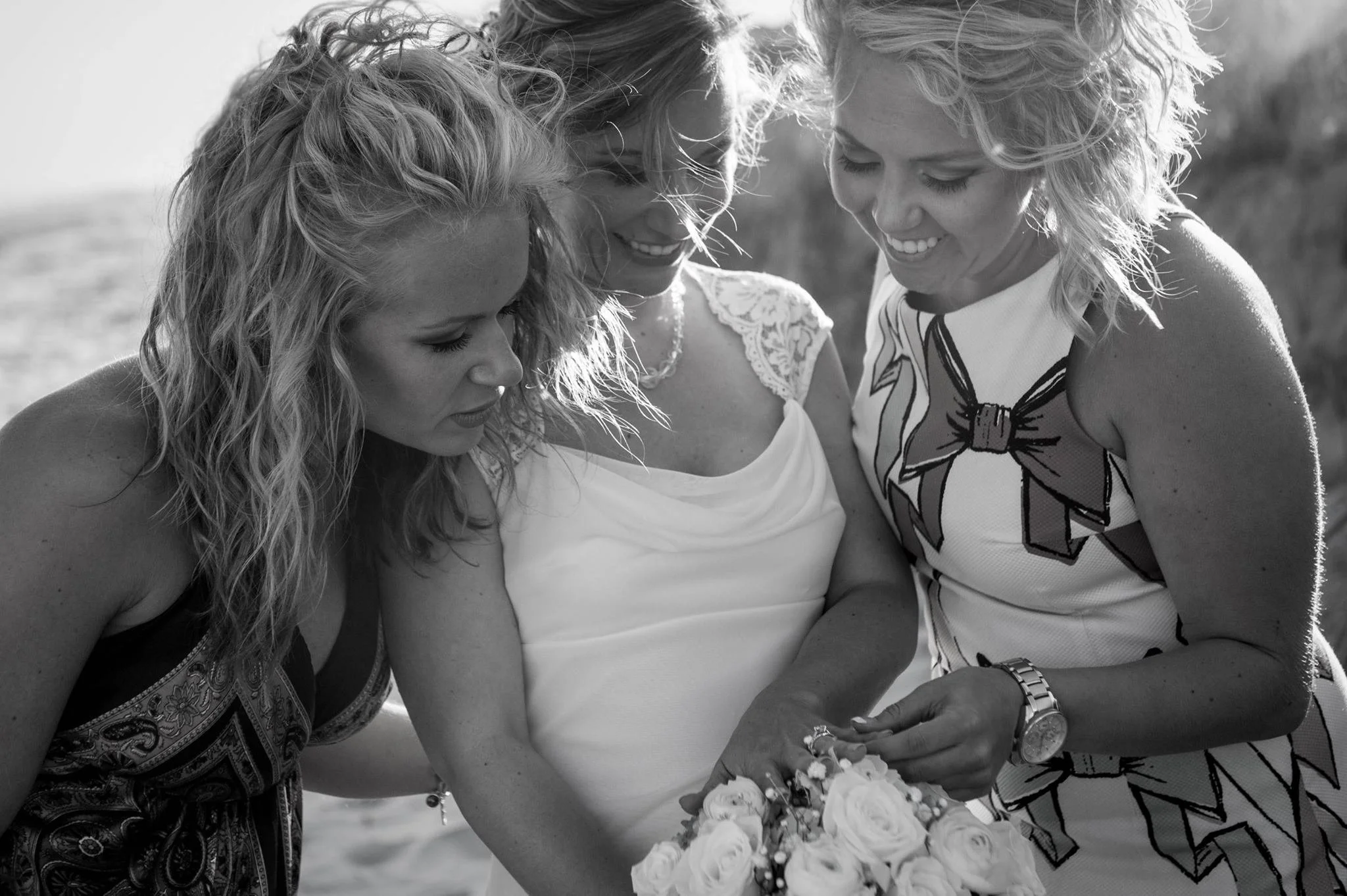 Three women smiling and looking down at a flower bouquet, outdoors.