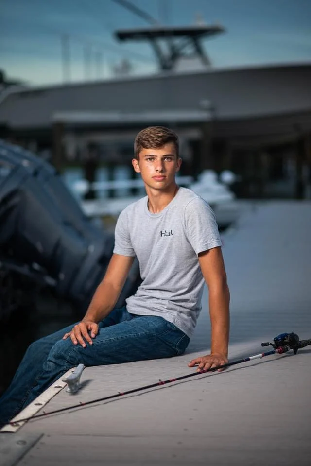 A teenage boy sitting on a dock next to a boat with a fishing rod on the deck, with a marina and boats in the background.