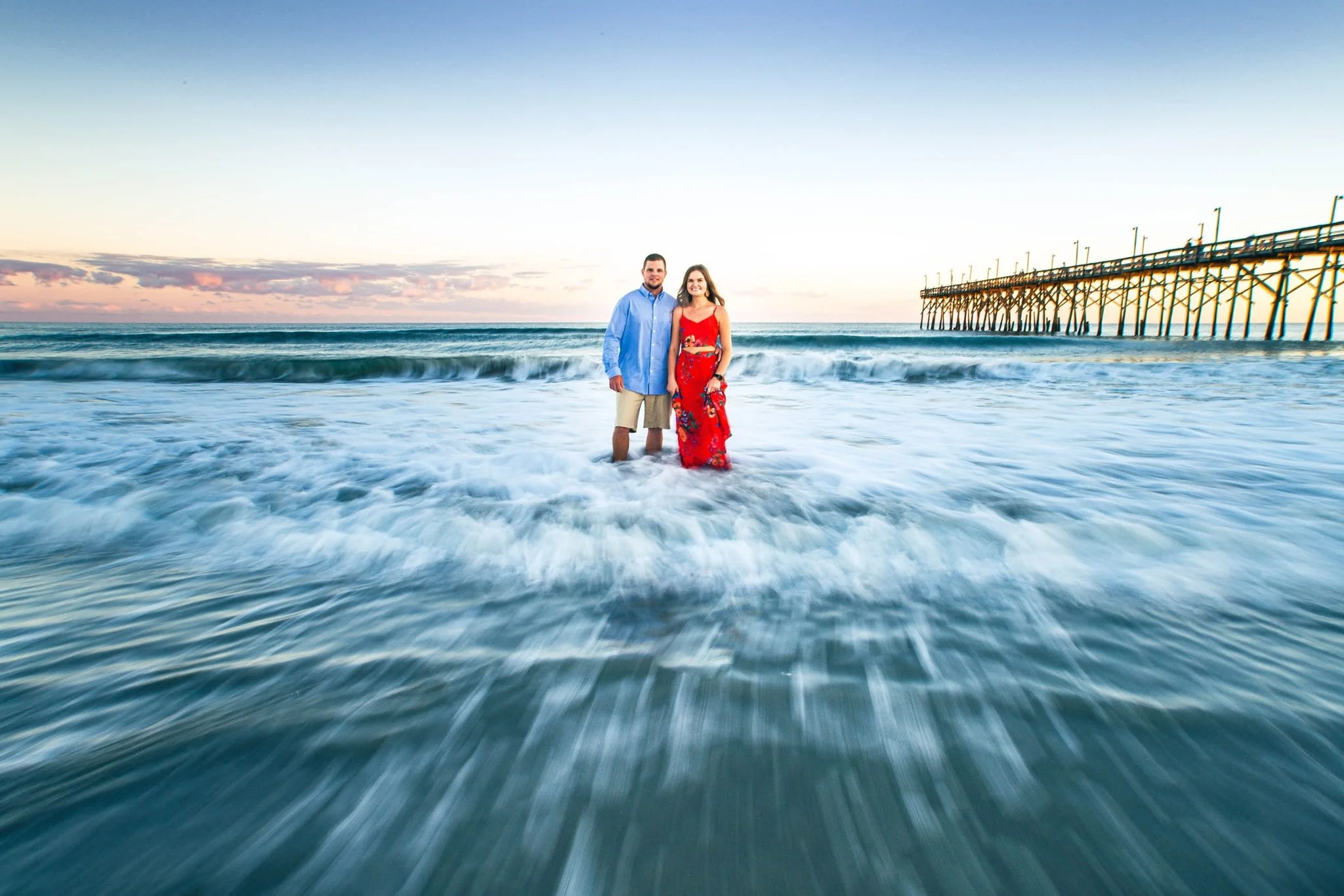 A couple standing in the shallow ocean waves with a pier in the background, during sunset.