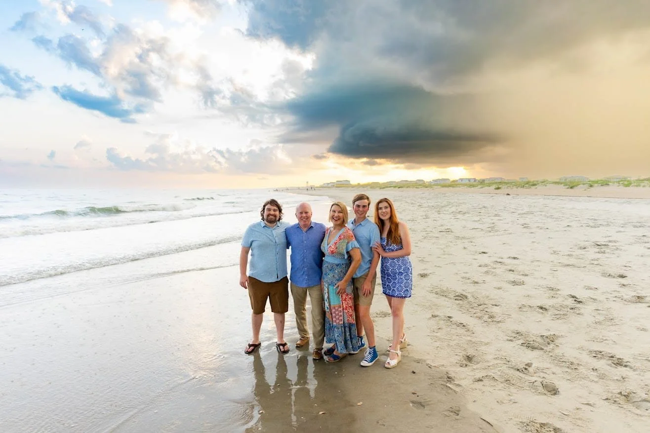 A group of five people standing on a beach with the ocean and a cloudy sky in the background during sunset.
