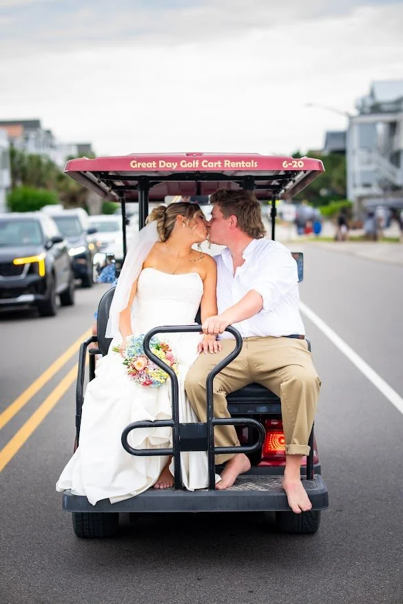 Bride and groom sitting on a golf cart, about to kiss, with the bride holding a bouquet, on an urban street during daylight.