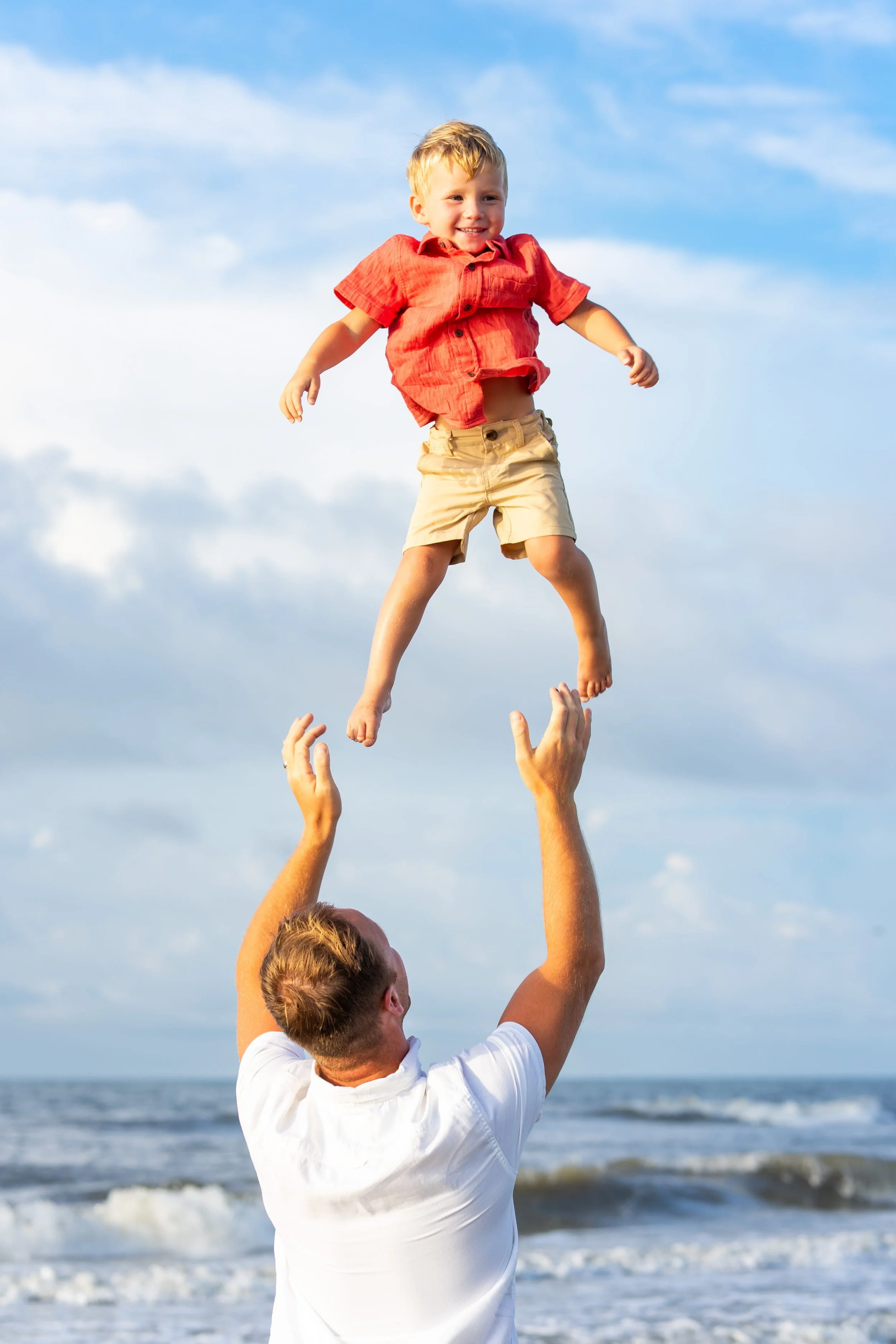 A man lifts a young boy into the air at the beach with ocean waves and a cloudy sky in the background.