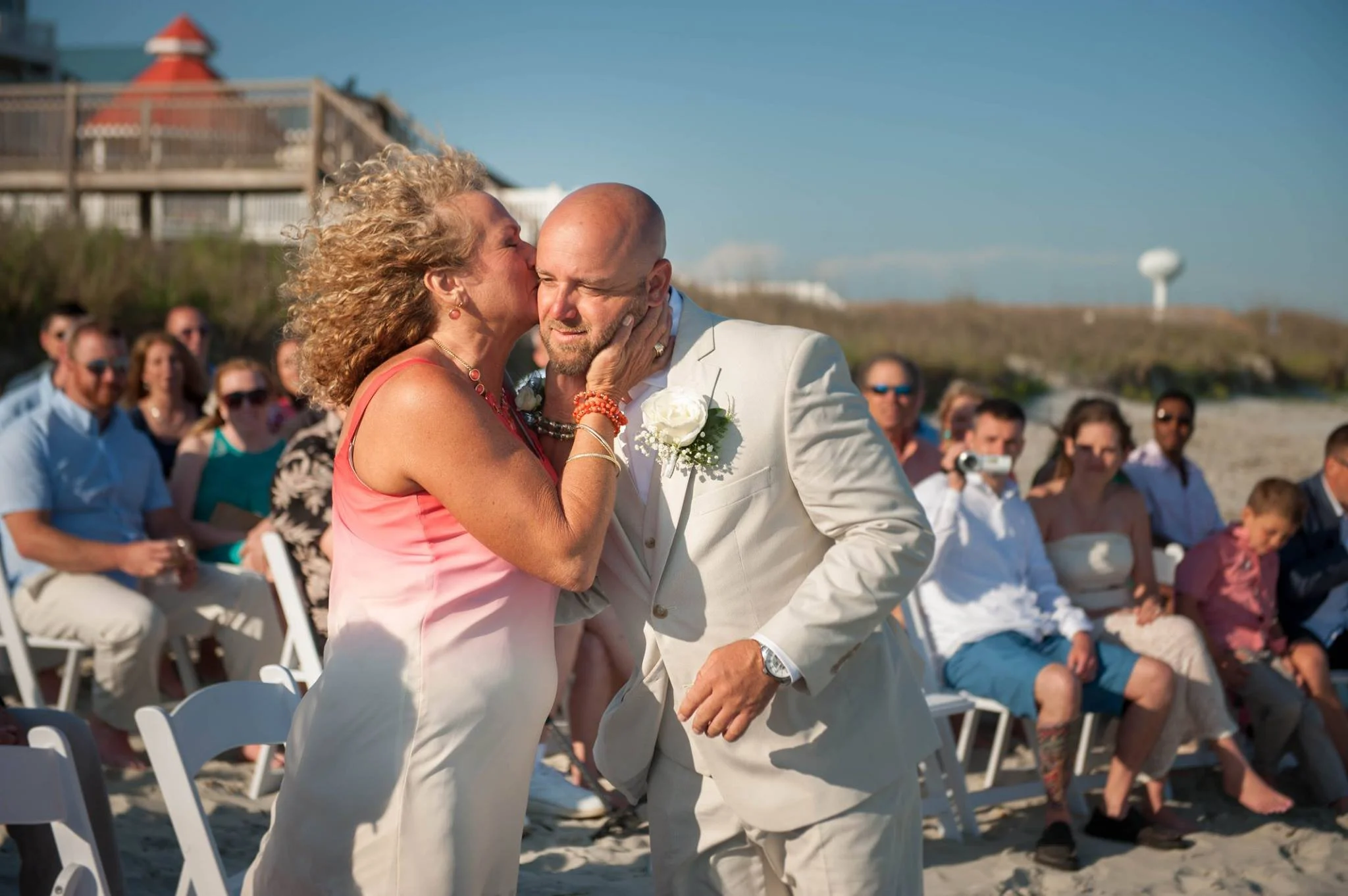 A woman kisses a man on the cheek at a beach wedding ceremony, with guests seated behind them and a water tower visible in the background.
