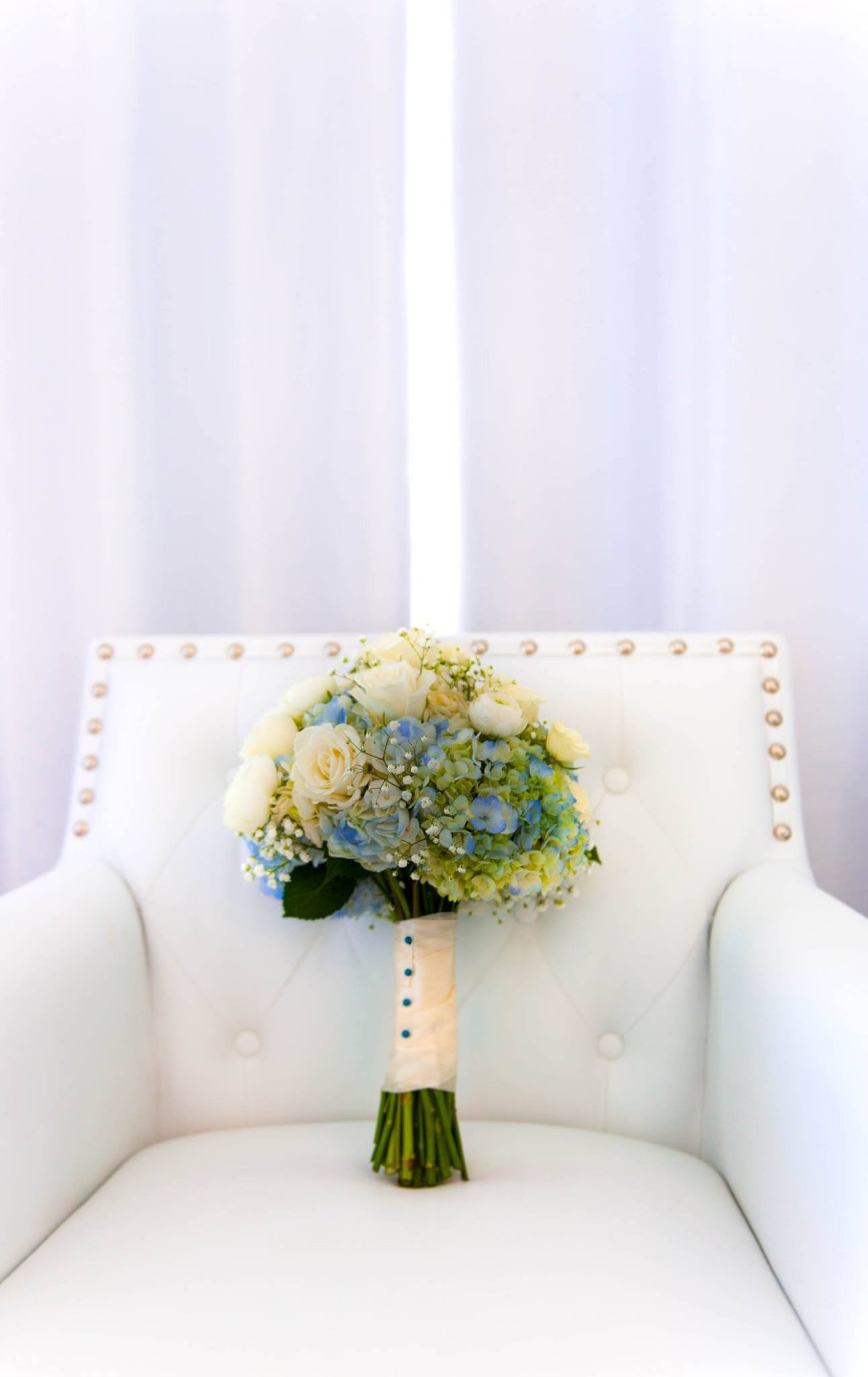 A bouquet of white and light blue flowers placed on a white cushioned chair with a tufted backrest and brass nailhead trim, against a plain white background.