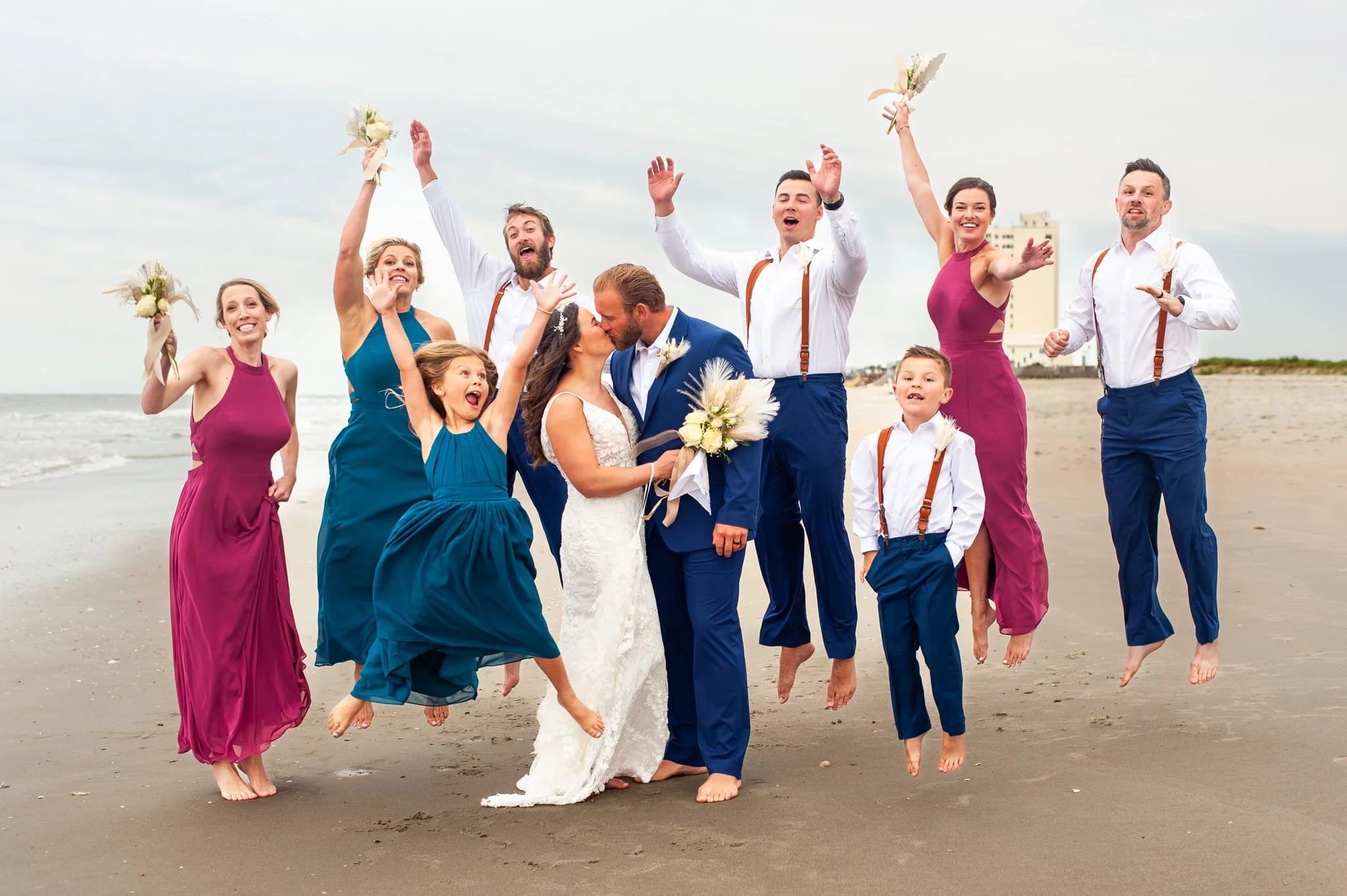 A wedding celebration on the beach with a bride and groom kissing surrounded by friends and family cheering and jumping.