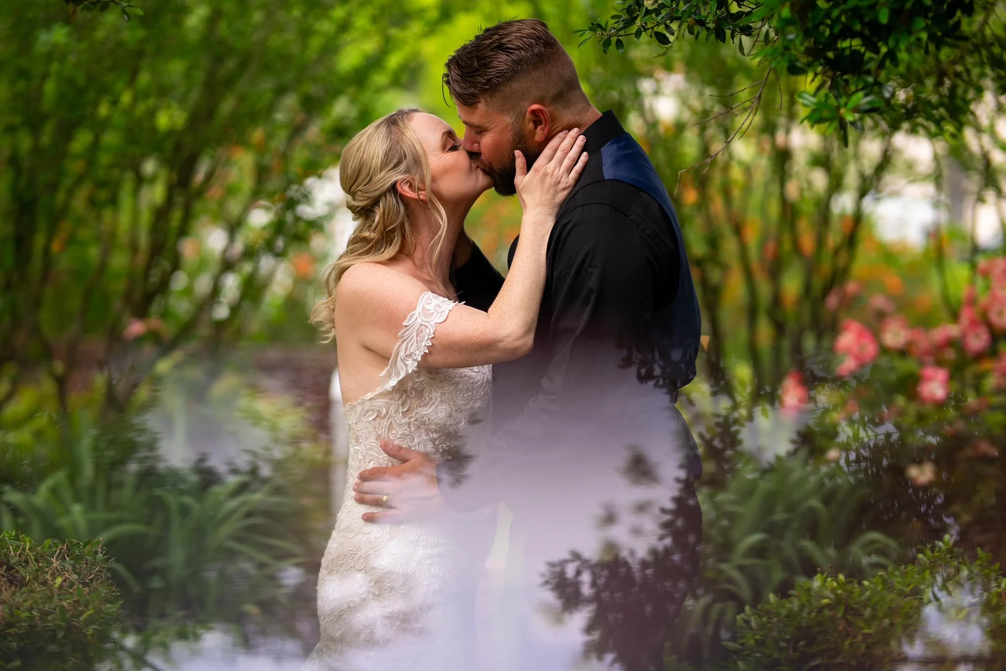 A couple sharing a kiss in a garden, with the woman in a lace wedding dress and the man in a black suit, surrounded by greenery and pink flowers.