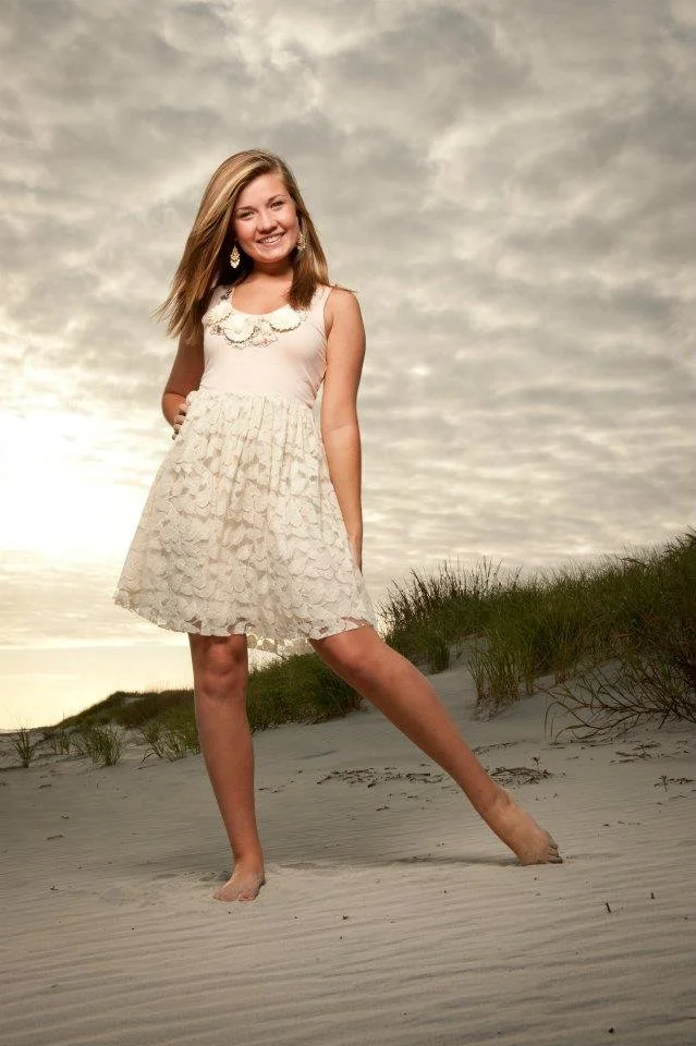 A young girl with long blond hair wearing a white dress with lace details standing barefoot on a sandy beach with patches of grass, smiling at the camera during sunset or sunrise.