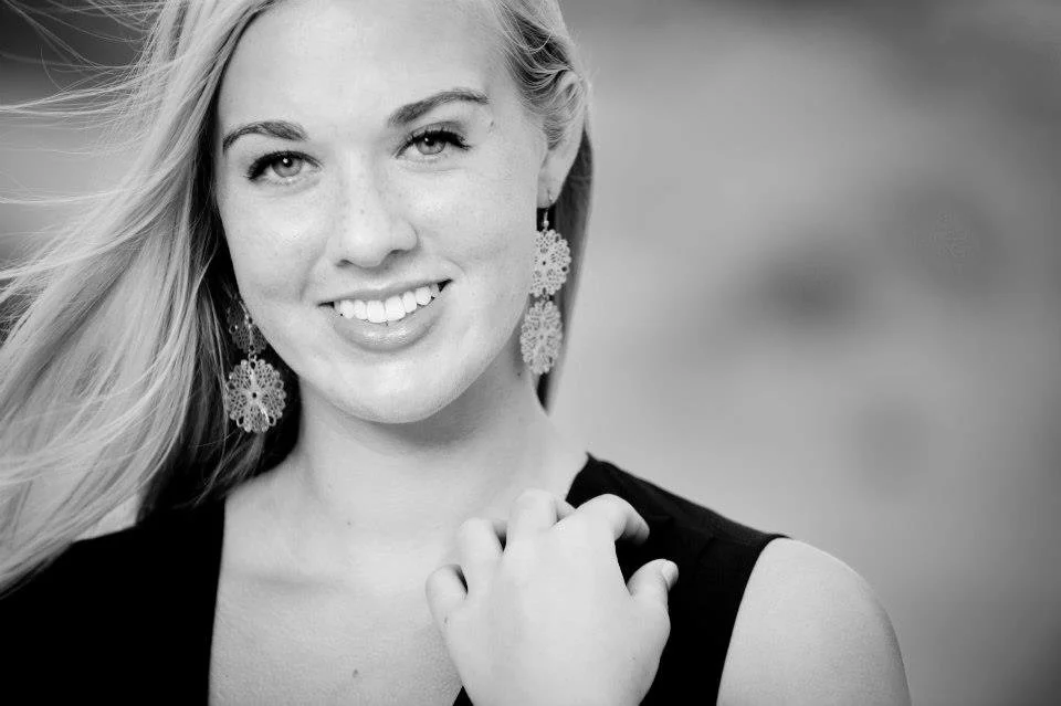 A smiling woman with long hair wearing floral earrings and a sleeveless top, touching her shoulder with her hand, in black and white.