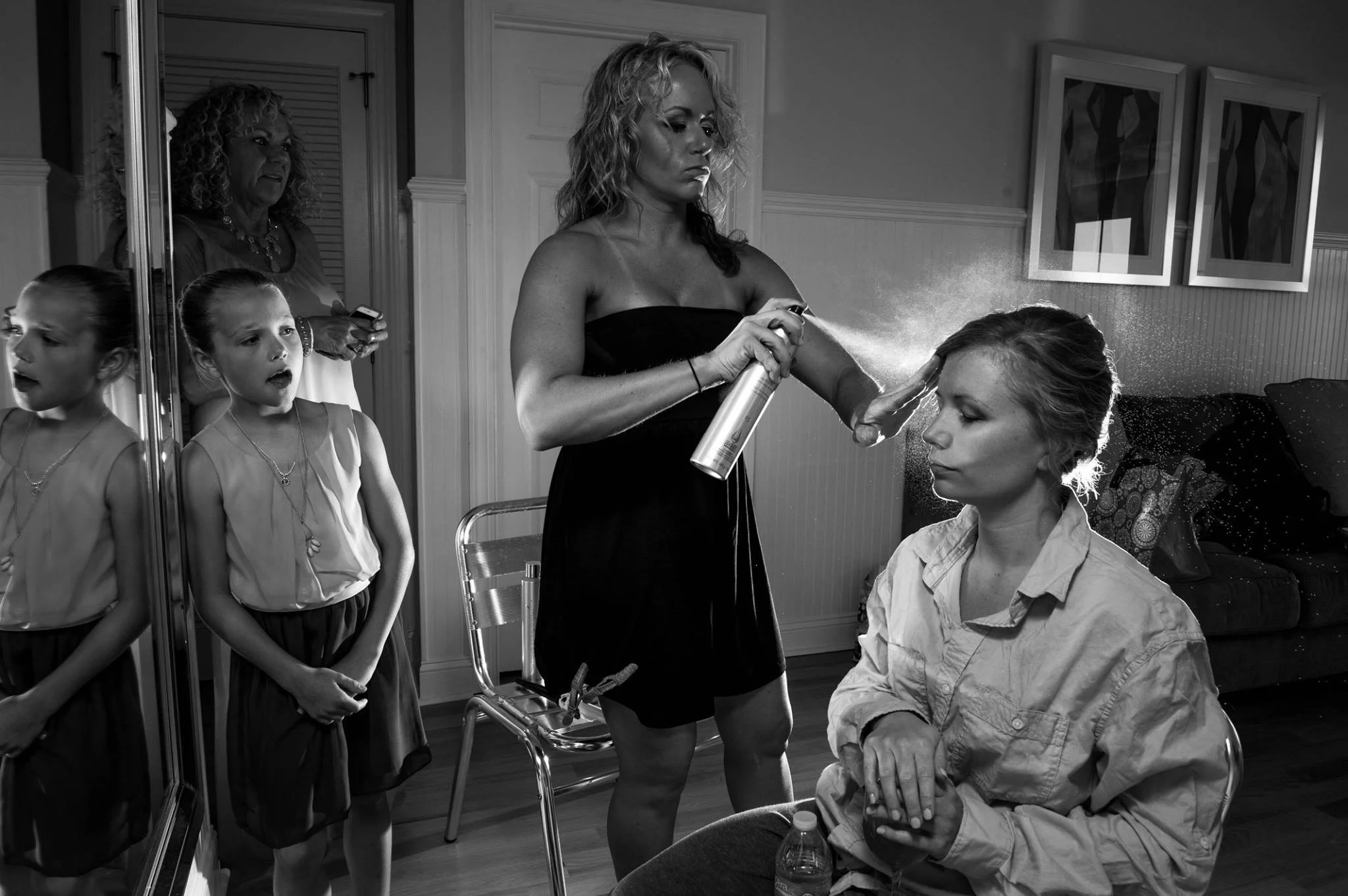 A woman is spraying hairspray on a seated woman’s hair in a room with two framed pictures on the wall. Two young girls and an older woman are standing nearby, watching, in front of a mirror.