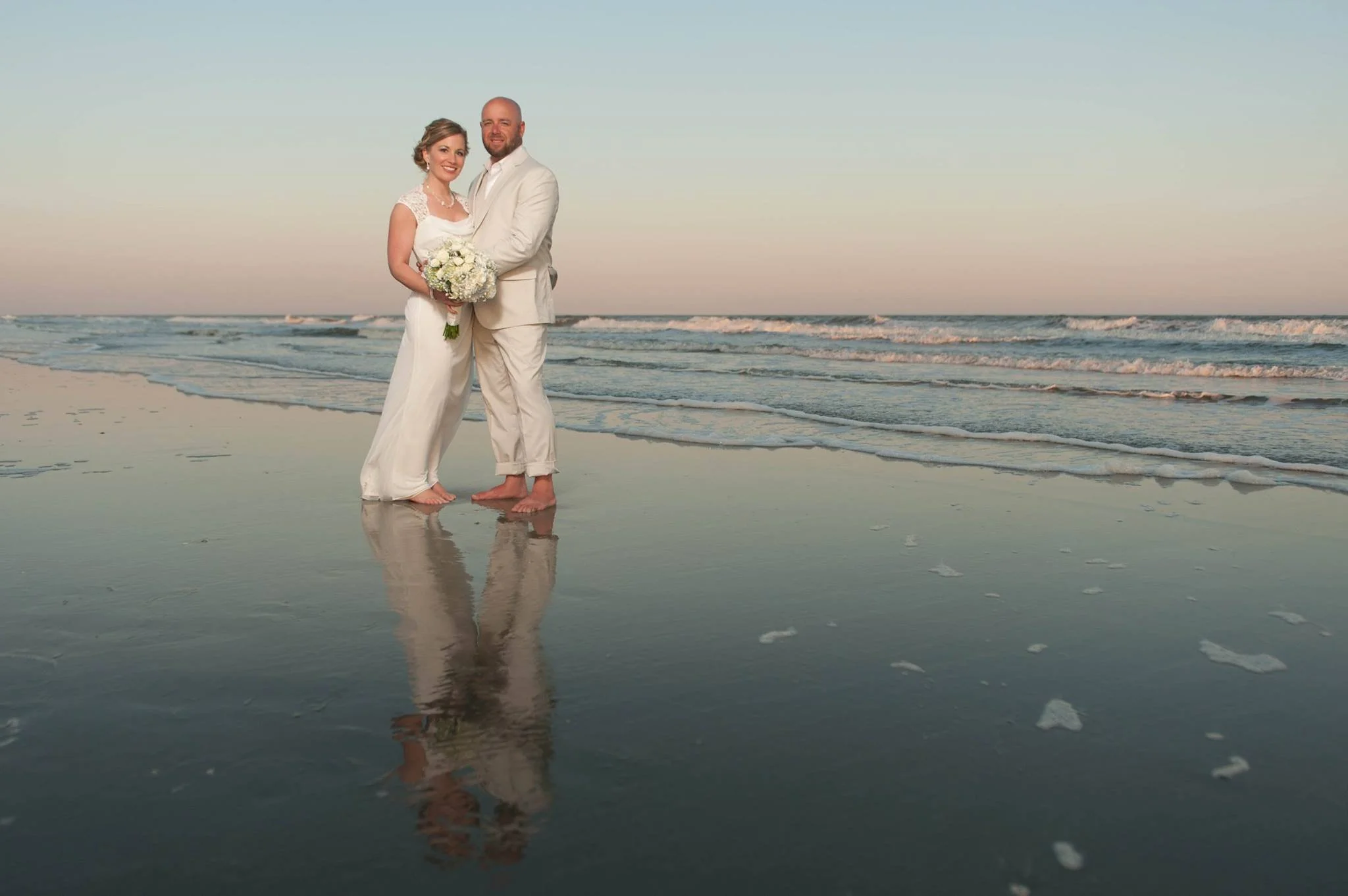 A newlywed couple standing on a beach at sunset, with the ocean and waves behind them. The bride is holding a bouquet of white flowers, and both are dressed in white wedding attire.