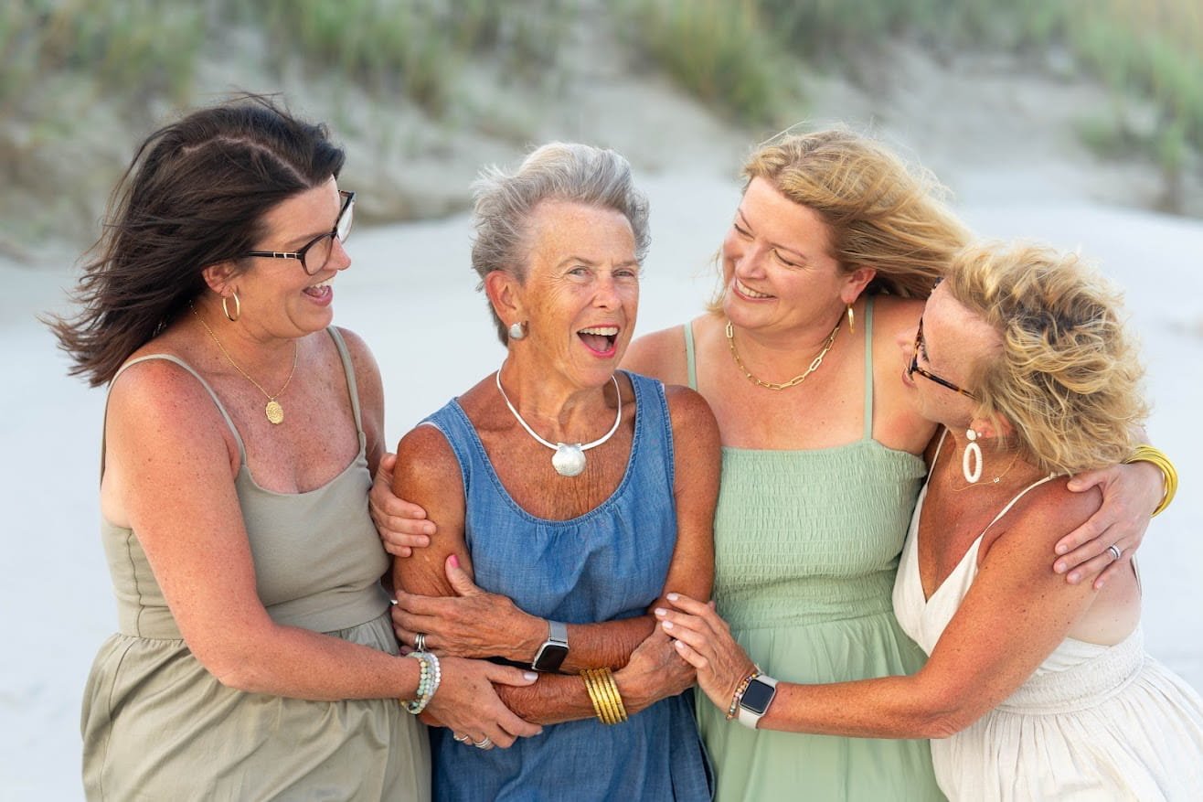 Four women, aged middle-aged to elderly, smiling and laughing together on a beach, embracing each other in a joyful group hug.