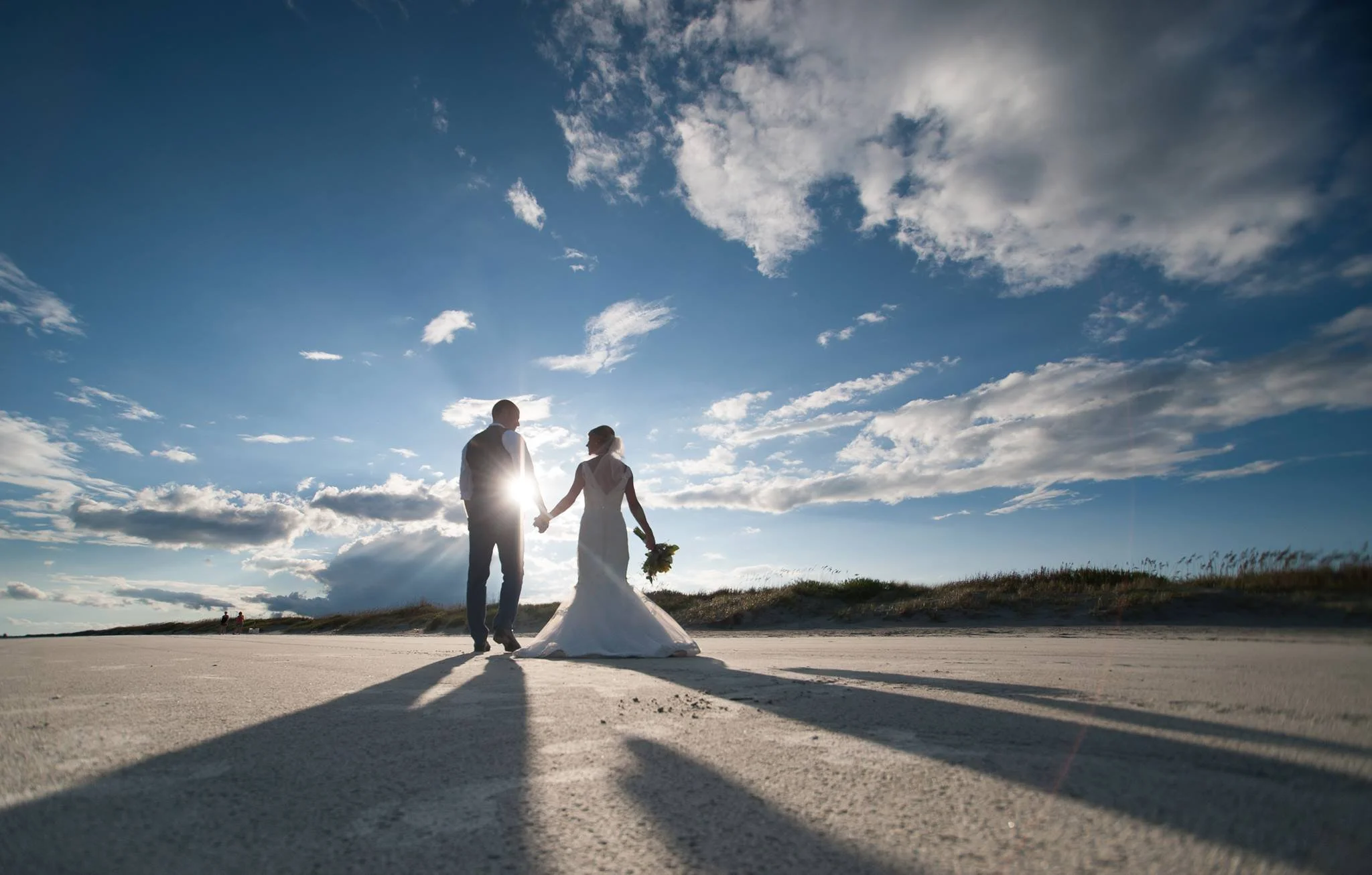 A bride and groom holding hands on a beach at sunset, with their shadows cast on the sand, and the sun shining behind them against a partly cloudy sky.