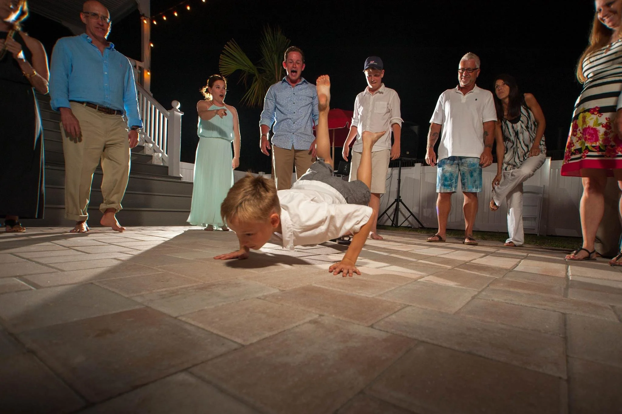 A young boy performing a push-up on the ground during a party at night, surrounded by adults and children watching and reacting, with some cheering and smiling, on a patio area with string lights and a fence.
