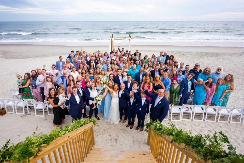 Large wedding group photo on the beach with a wedding arch in the background, guests in formal attire, ocean waves, sandy shore, and clear sky.
