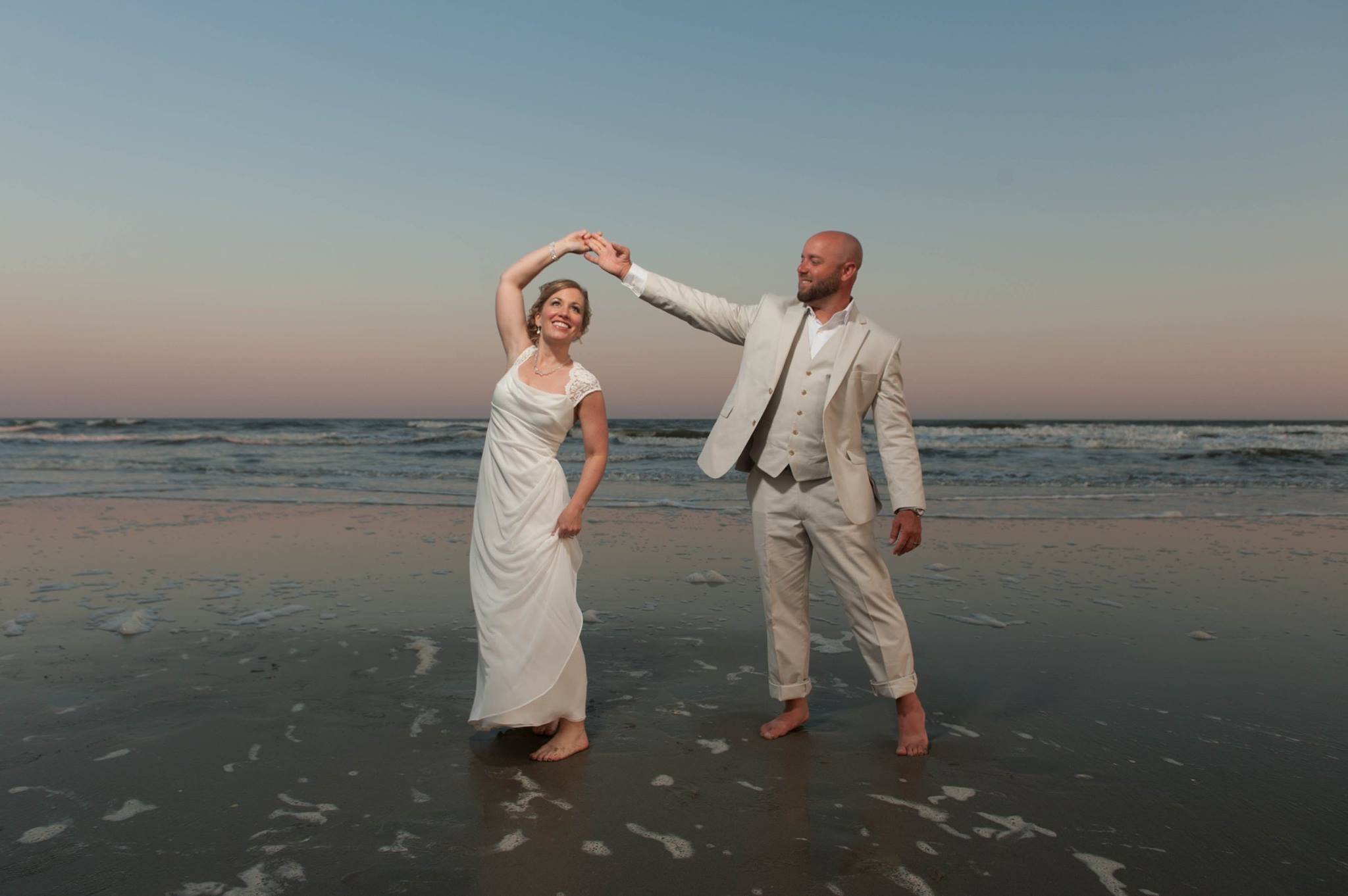 A couple in wedding attire dancing on the beach at sunset or dusk with the ocean in the background.