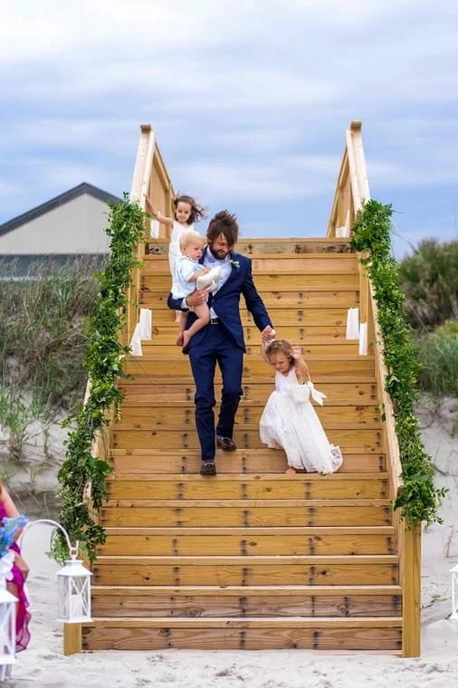 A man and three young girls descending a wooden staircase decorated with greenery outdoors, with a house and sky in the background.