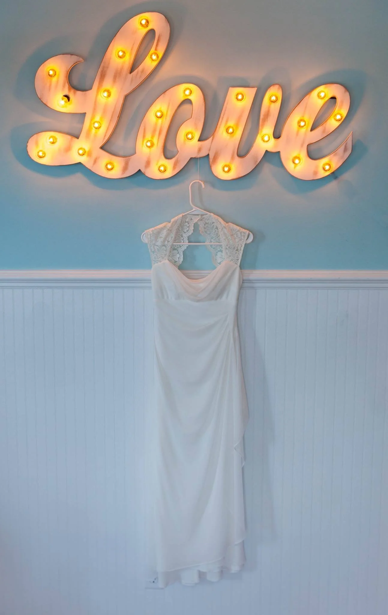 A wedding dress hanging on a wall with a large illuminated marquee sign spelling 'Love' above it.