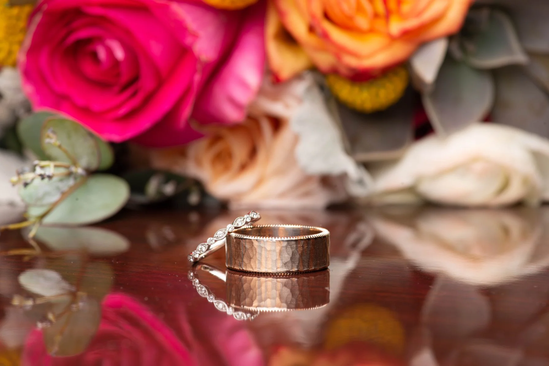 A pair of wedding rings, one with diamonds, placed on a reflective wooden surface with a bouquet of pink, peach, and white roses and eucalyptus leaves in the background.