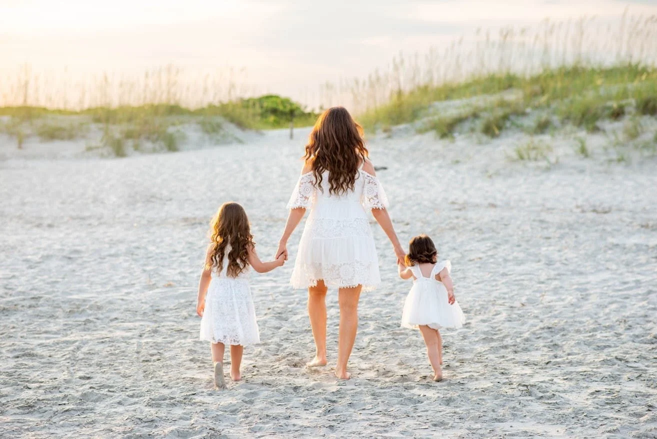 A woman and two young girls walking on a sandy beach, holding hands, all dressed in white dresses. The scene appears to be at sunset or sunrise with soft, golden lighting.
