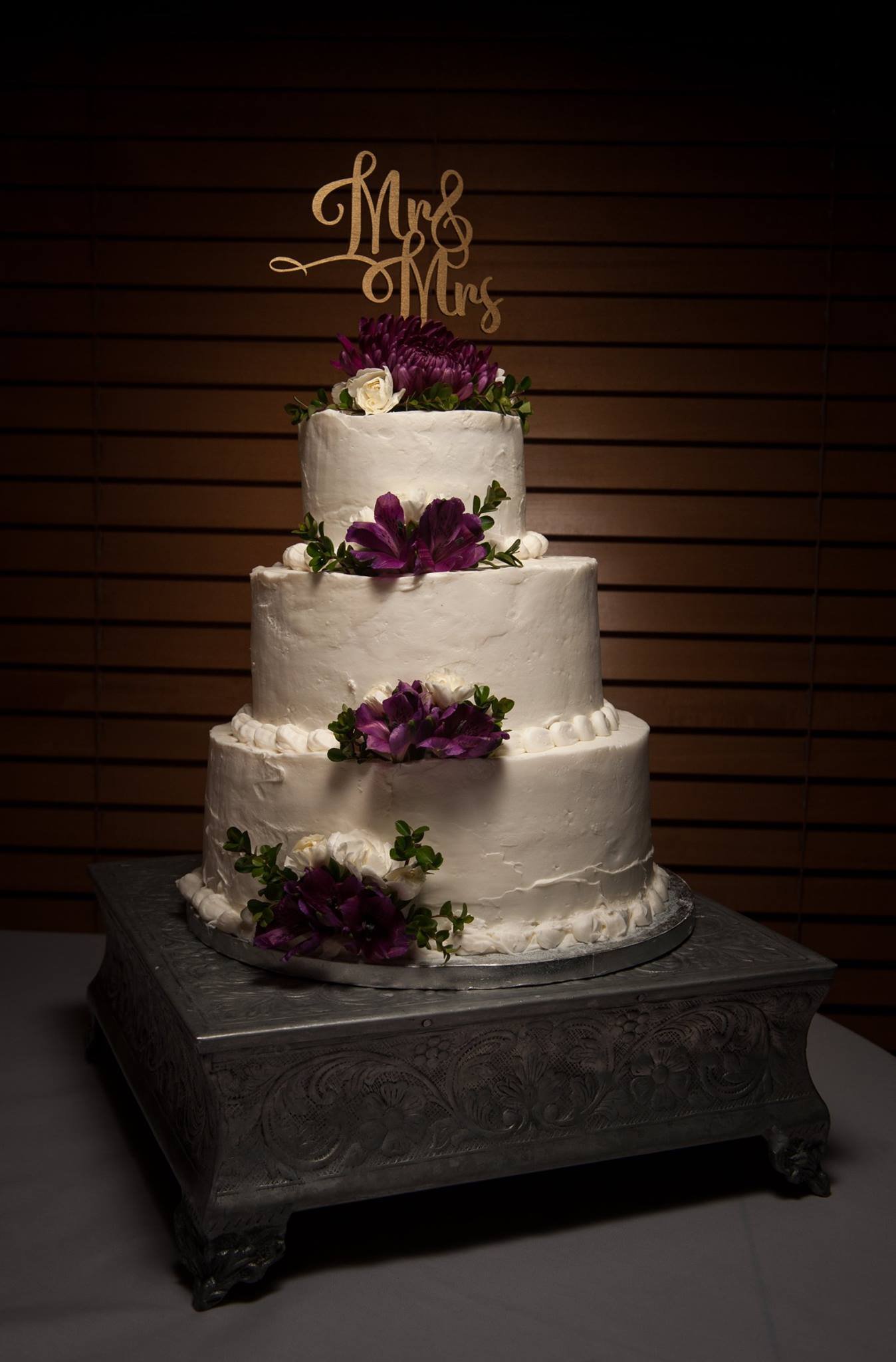 A three-tier white wedding cake decorated with purple and white flowers, with a "Mrs & Mrs" cake topper, on a silver ornate stand.