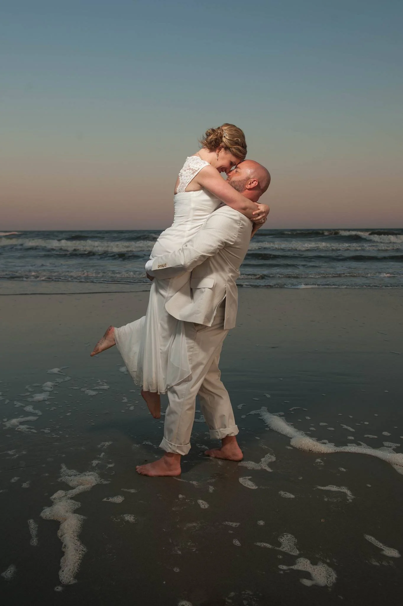 A couple in wedding attire on the beach, with the man lifting the woman in his arms as they stand barefoot in the sand near the water, at sunset.
