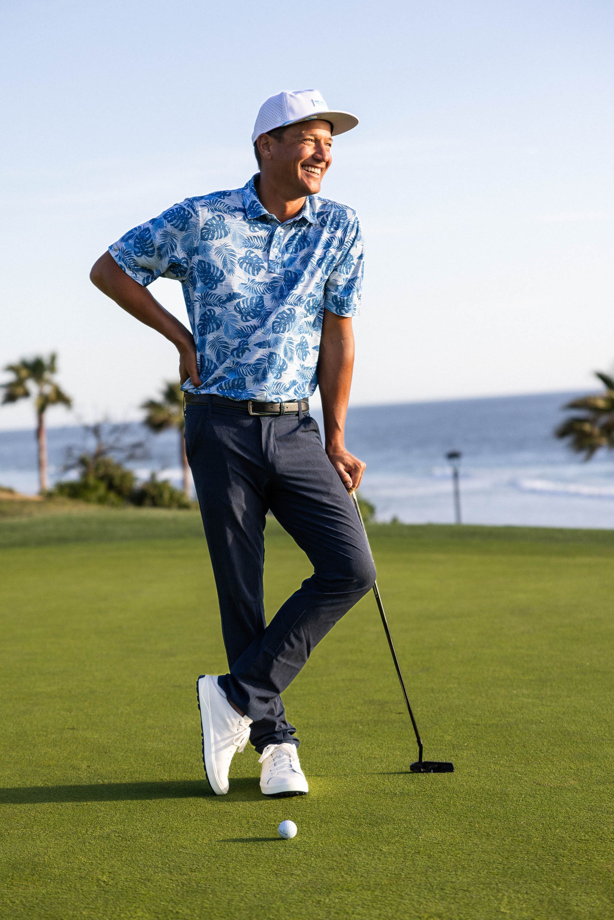 A man in a blue and white patterned shirt, dark pants, and a white cap stands on a golf course holding a golf club behind a golf ball on the green. OluKai Golf campaign photographed by Scott Wild.
