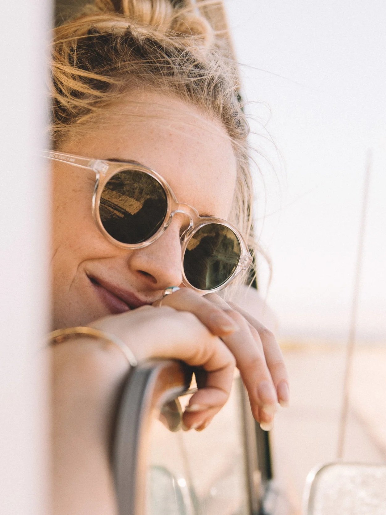 A woman with blonde hair and round sunglasses resting her head against a wall, smiling softly and looking down.
