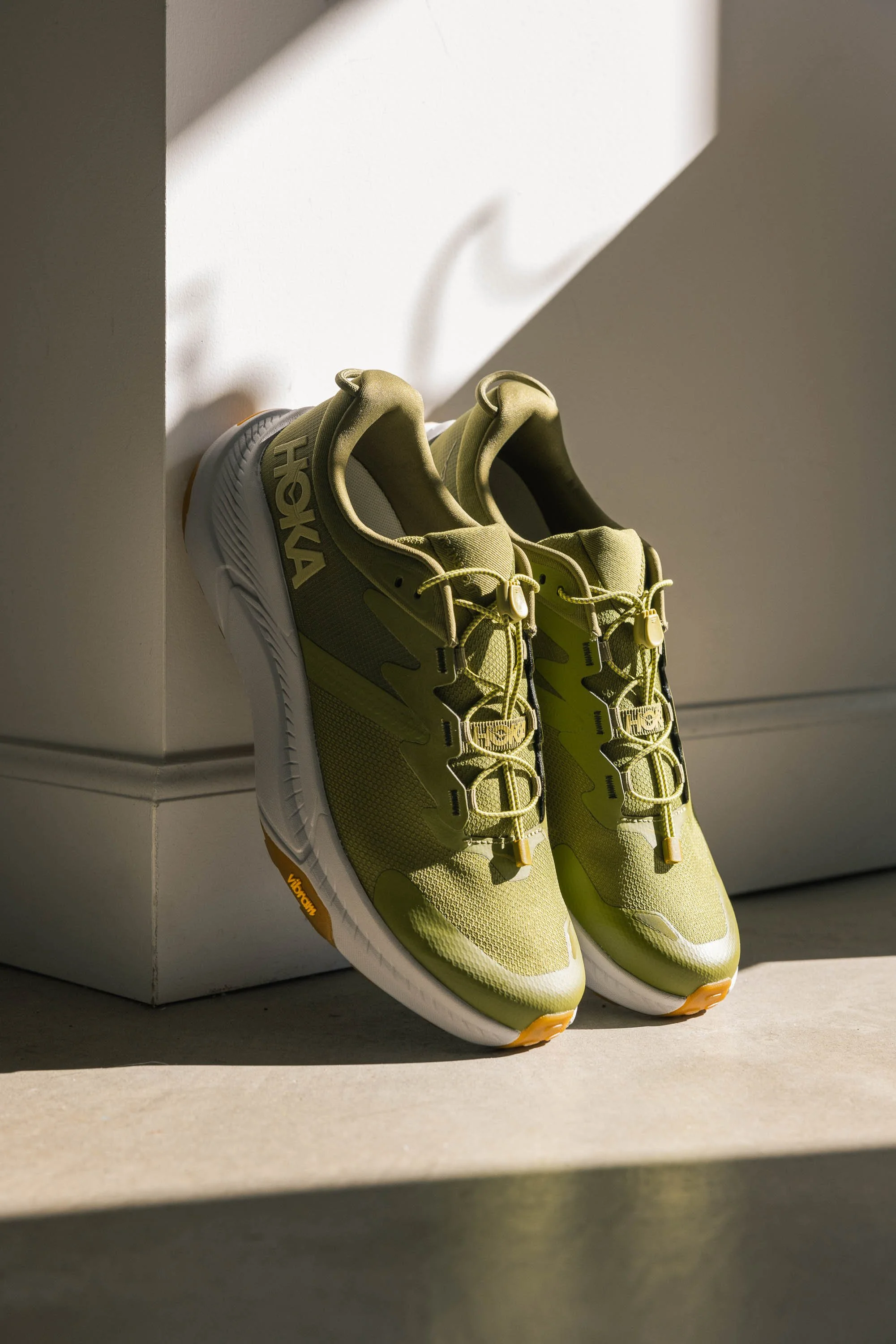 A pair of yellow Reebok athletic shoes placed on the floor, with sunlight casting shadows on the wall and part of the shoes. HOKA campaign photographed by Scott Wild.