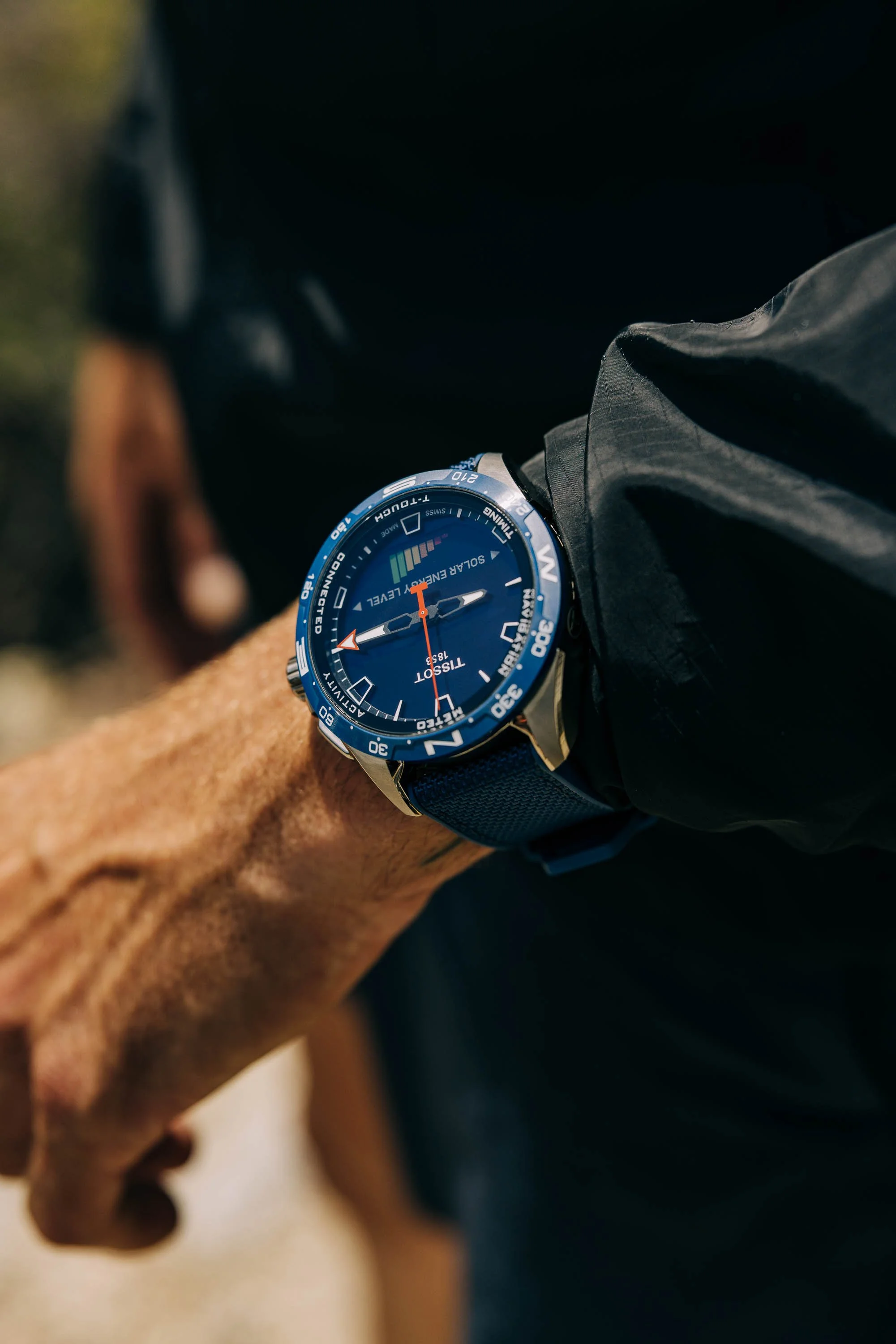 Close-up of a person wearing a blue wristwatch with a digital display, black wristband, and black jacket. Part of the person's arm and hand are visible. The background is blurred. TISSOT campaign photographed by Scott Wild.