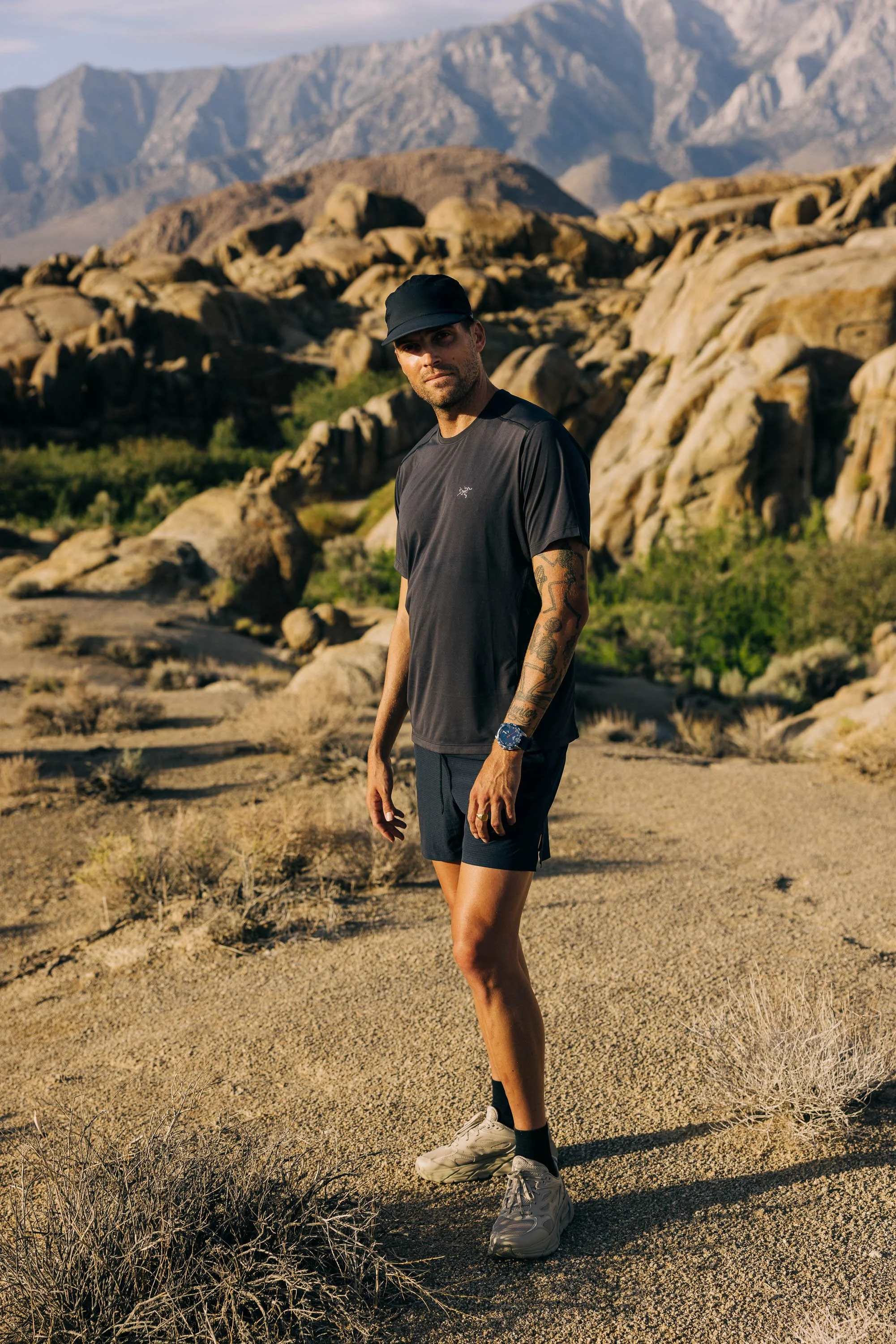 A man wearing a black cap, black t-shirt, black shorts, and sneakers stands in a desert landscape with rocky terrain and sparse desert vegetation, with mountains in the background. TISSOT campaign photographed by Scott Wild.