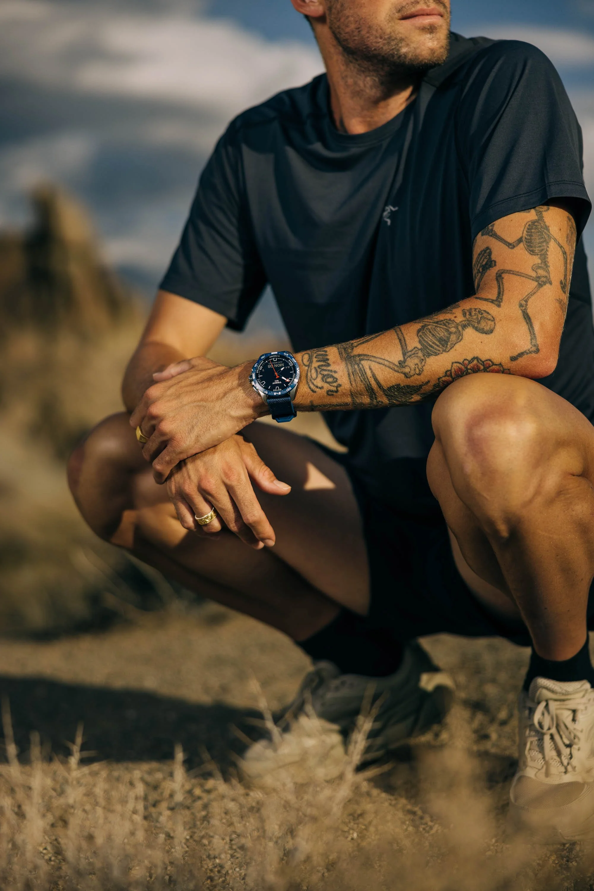 A man with a tattooed arm is sitting outdoors on a dirt ground, wearing a black t-shirt, gray sneakers, and a black watch, with a cloudy sky in the background. TISSOT campaign photographed by Scott Wild.