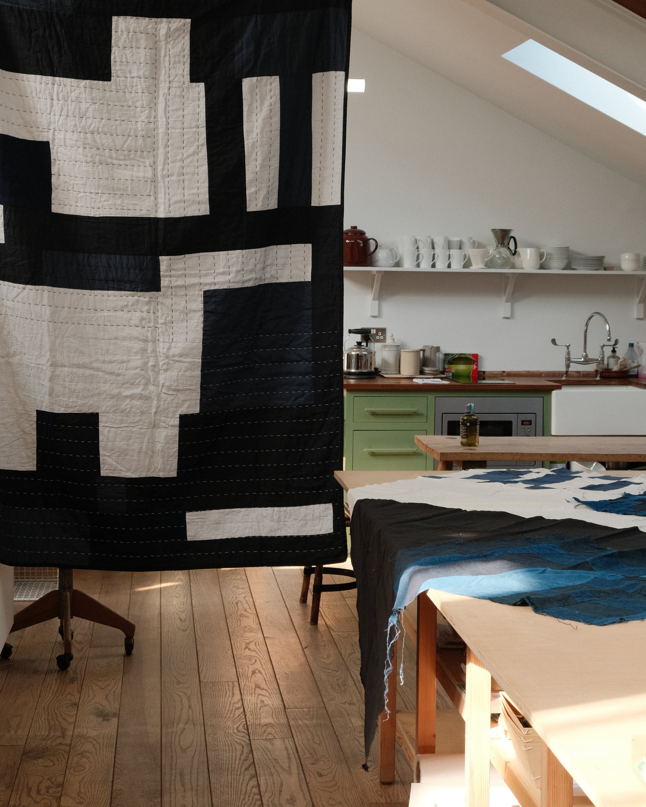 Part of a room with a large black and white quilt hanging, a green kitchen counter with various kitchen items, a wooden table with fabric, and wooden flooring.