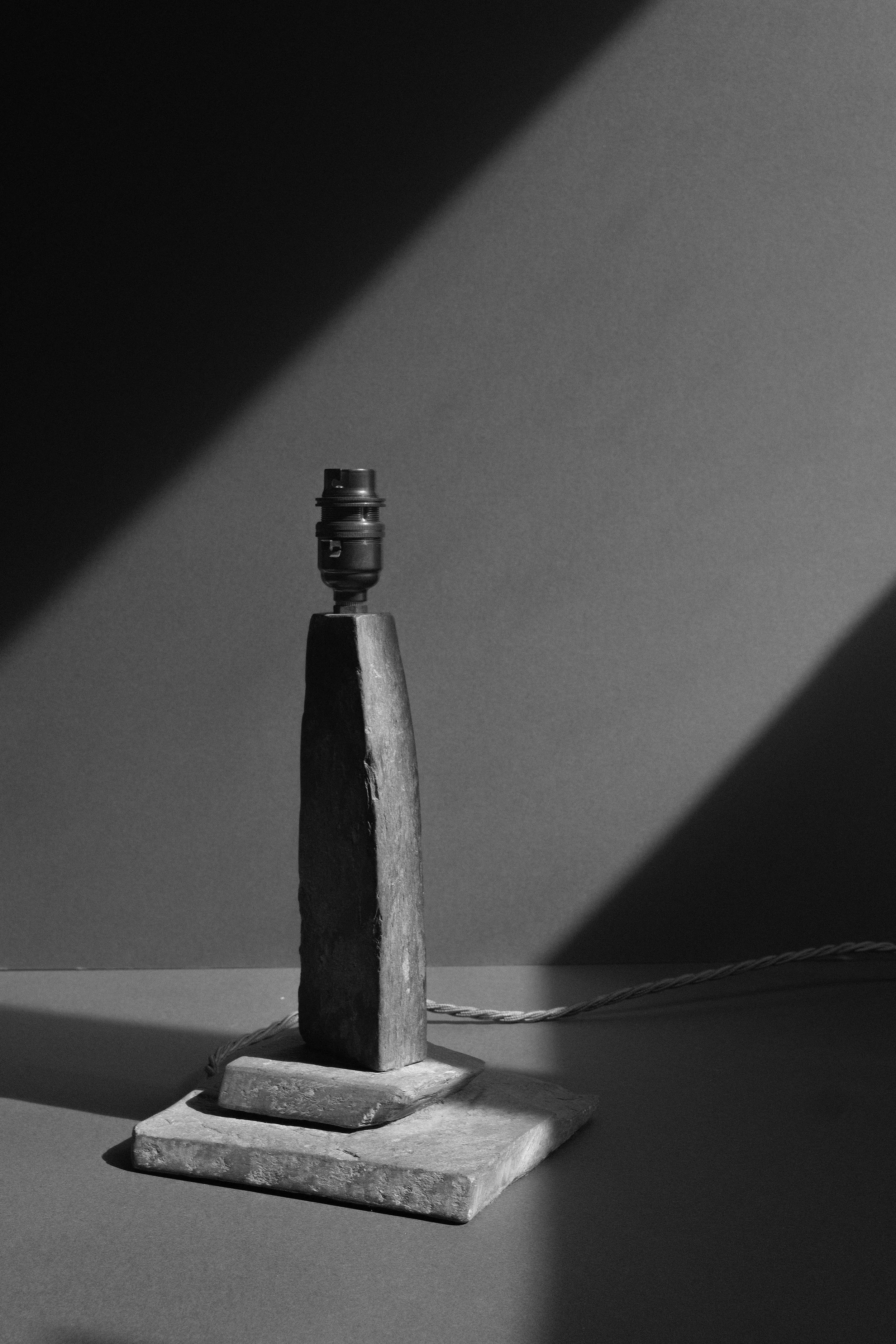 A black and white photo of a small lamp with a stone base and a wooden middle section, with a cord extending to the right. The lamp is turned off and is illuminated by light coming from the left side.